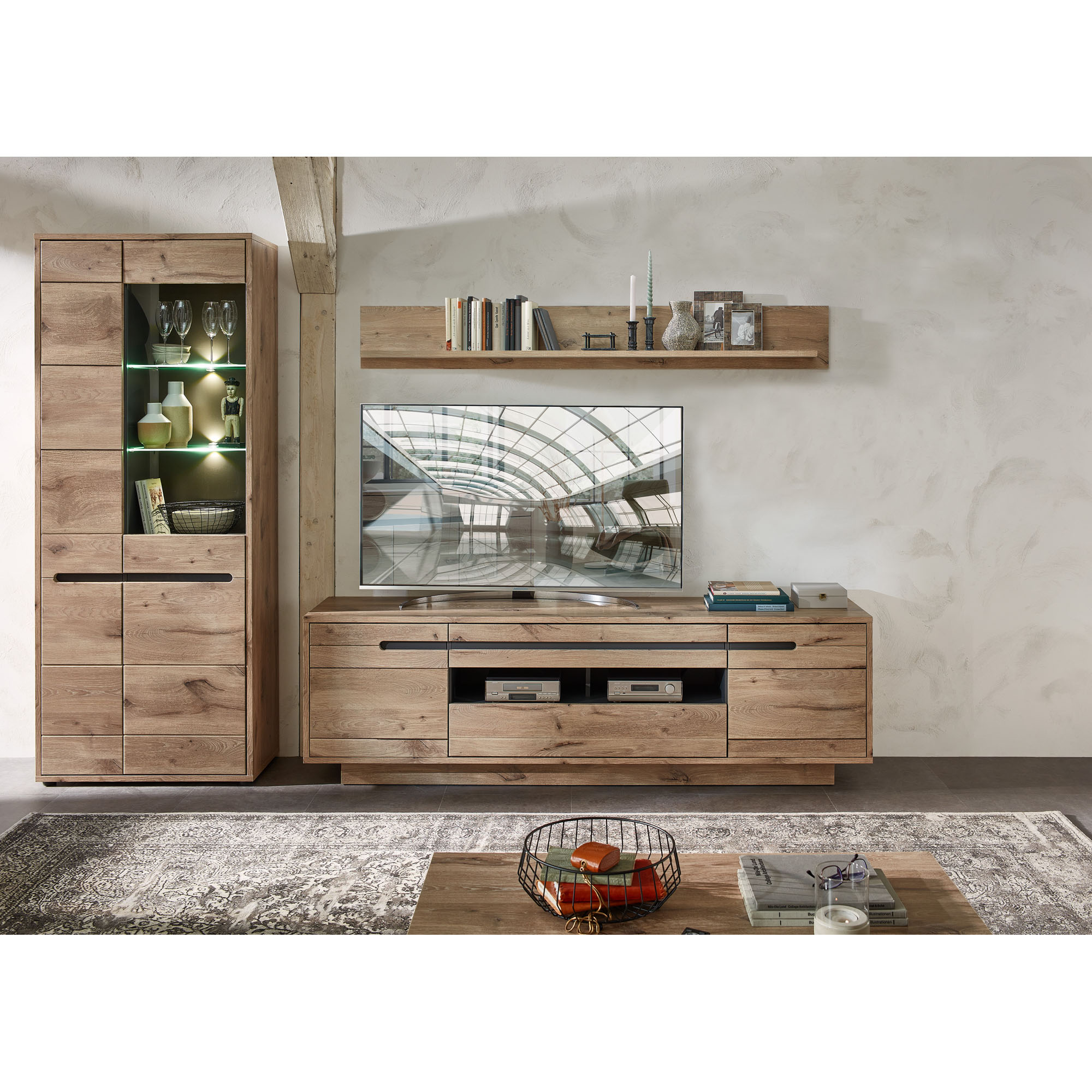 Ein Wohnzimmer mit einer Vitrine links, einem TV-Lowboard in der Mitte und einem Wandboard darüber. Ein Fernseher steht auf dem Lowboard, davor liegt ein Teppich und ein Couchtisch.