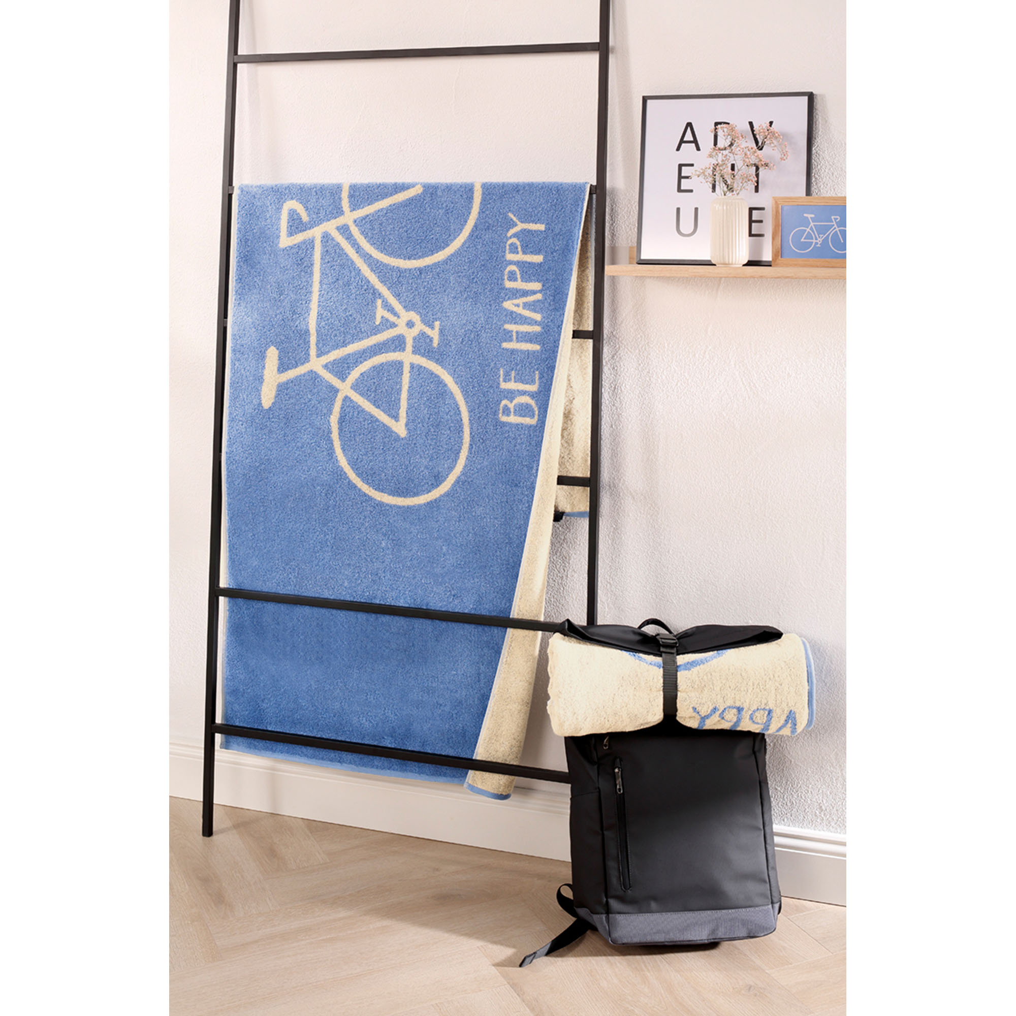 Blaues Travel-Strandtuch mit Fahrradmotiv hängt über einer schwarzen Leiter in einer hellen Raumszene.