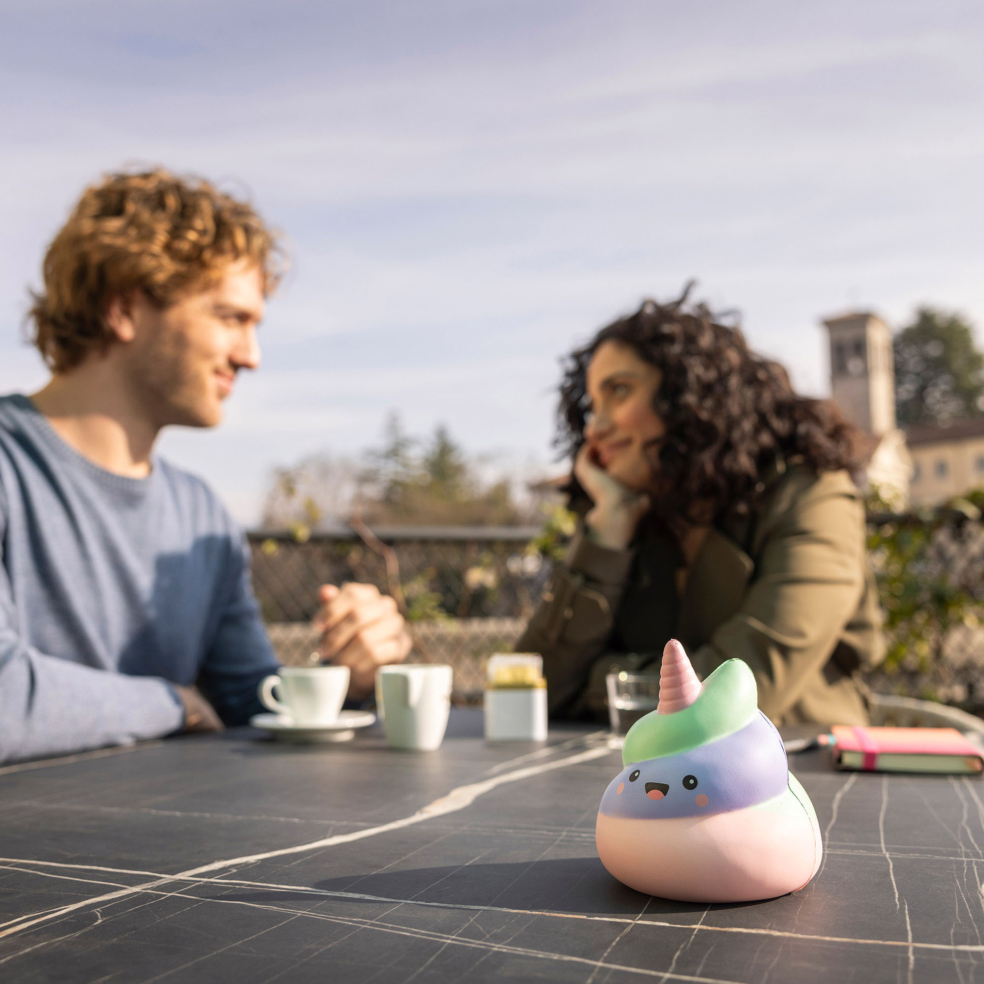 Pastellfarbene Antistress-Figur in Häufchenform mit Einhornhorn, auf einem dunklen Tisch vor zwei sitzenden Personen dargestellt.