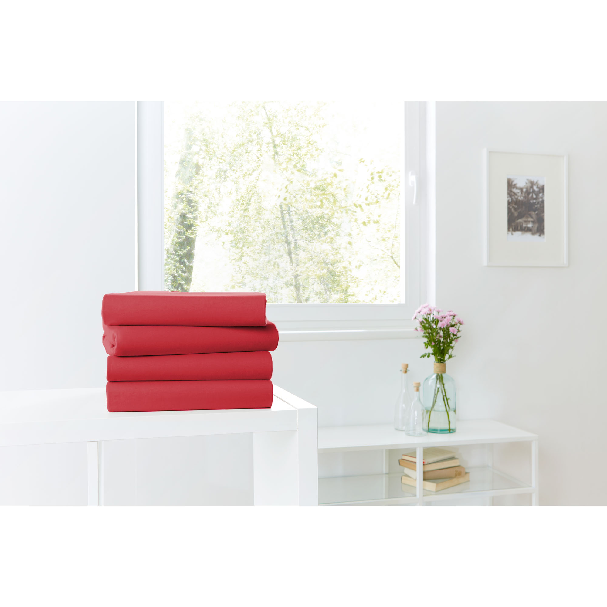 Vier gefaltete Spannbetttücher in Rot auf einer weißen Kommode vor einem hellen Fenster; Größe 100 x 200 x 30 cm.