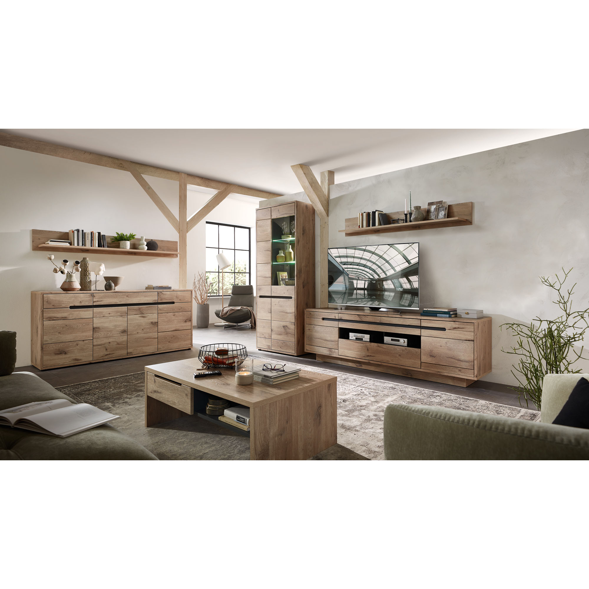 Ein Wohnzimmer mit mehreren Möbelstücken in Holzoptik. Über dem TV-Lowboard hängt ein Wandboard, darauf stehen Bücher und Dekoobjekte. Weitere Möbel wie Vitrine, Sideboard und Couchtisch sind sichtbar.