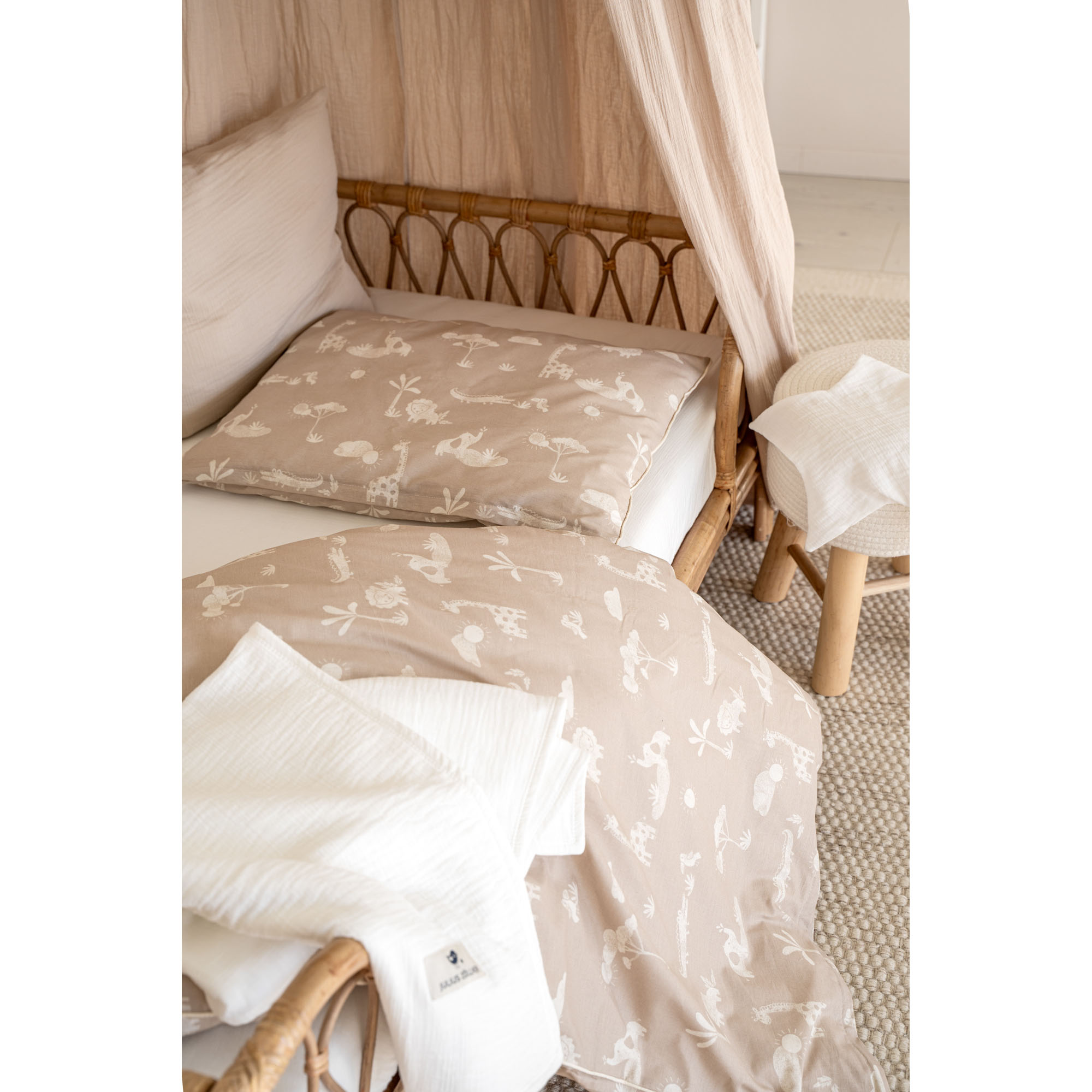 Beigefarbene Kinderbettwäsche mit weißen Safari-Tiermotiven auf einem Rattanbett mit cremefarbenem Baldachin und weißen Textilien