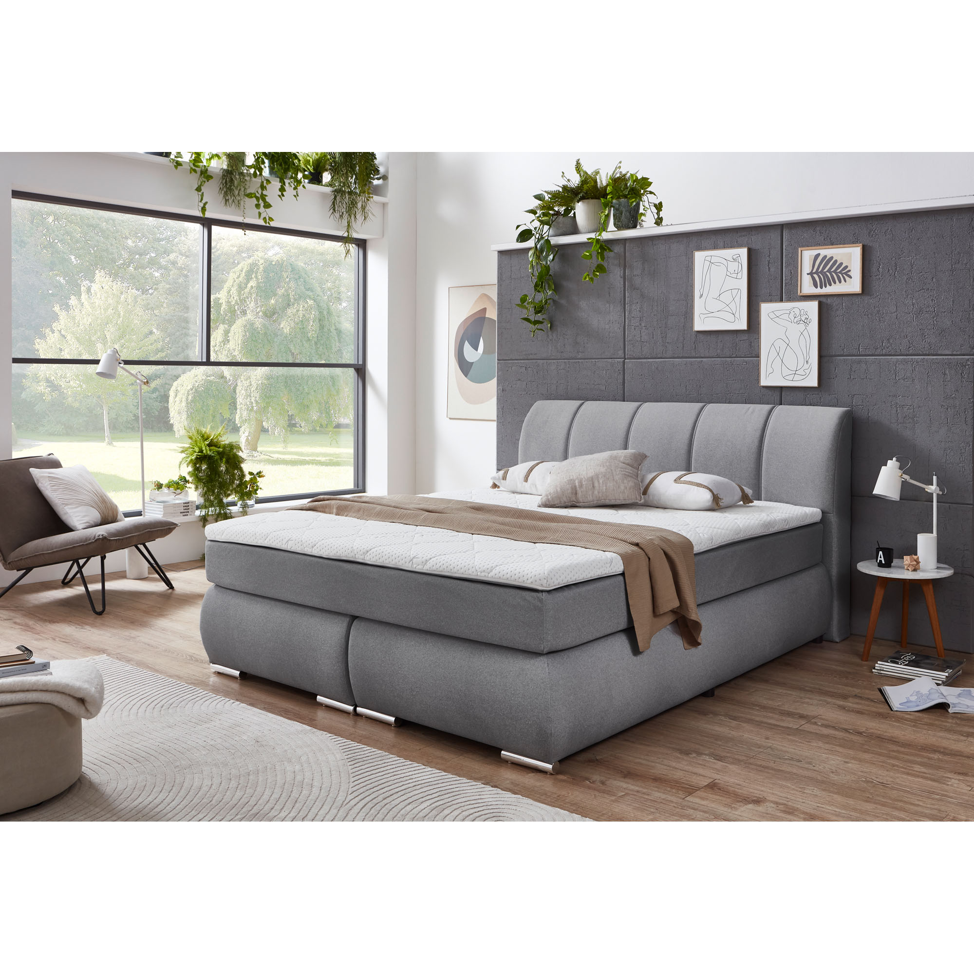 Graues Polsterbett mit gepolstertem Kopfteil in einem Schlafzimmer mit großer Fensterfront, Pflanzen und Wandbildern