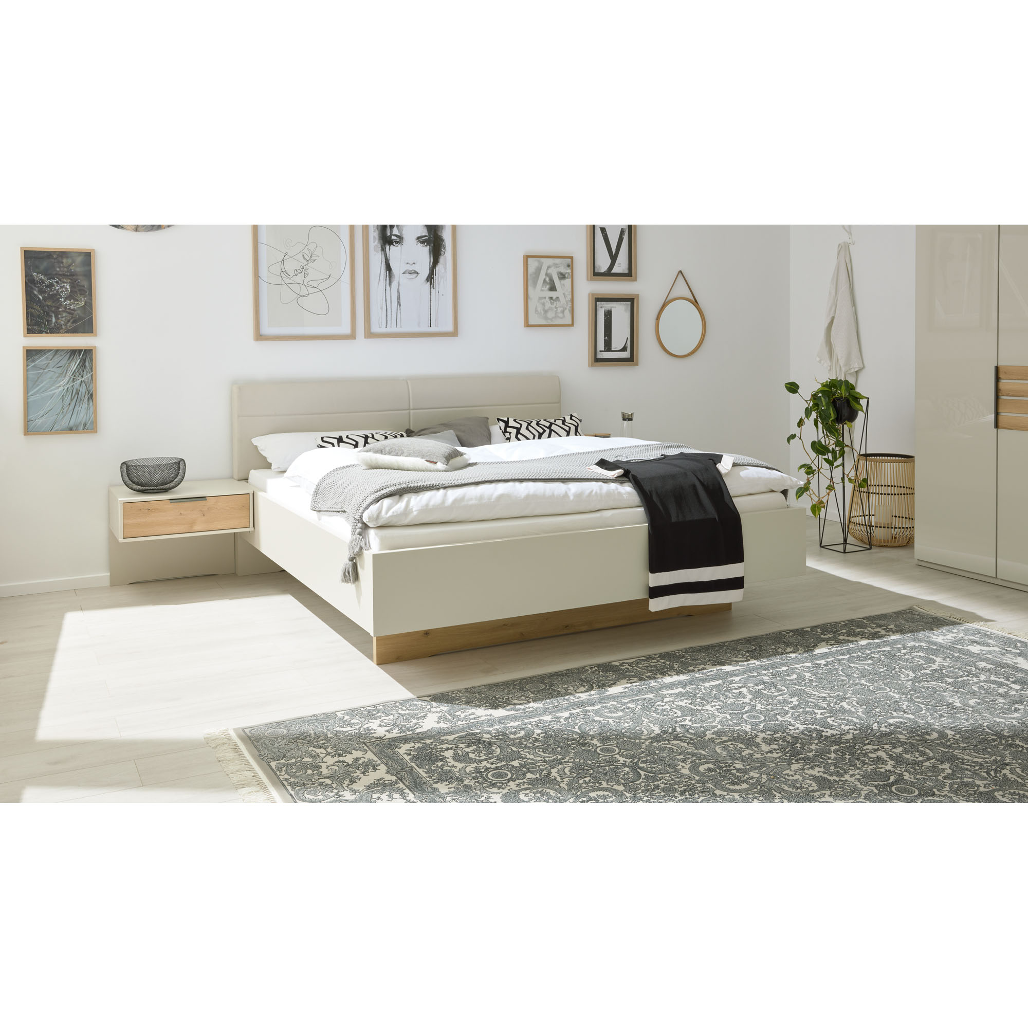 Helles Doppelbett Interliving 1026 mit gepolstertem Kopfteil in einem modern eingerichteten Schlafzimmer mit Nachttisch, Wandbildern und Teppich