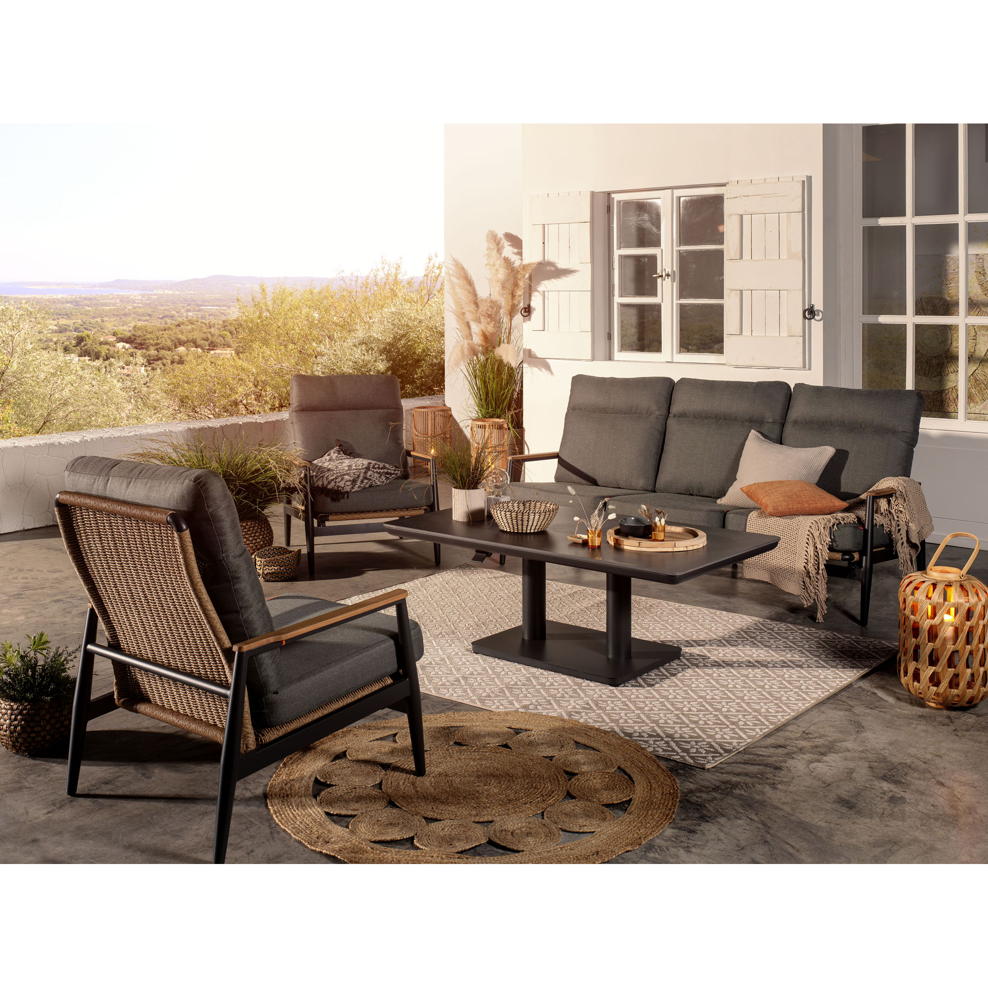 Ein Sofa und mehrere Sessel mit grauen Polstern stehen um einen rechteckigen Tisch auf einer überdachten Terrasse mit Ausblick in die Landschaft.