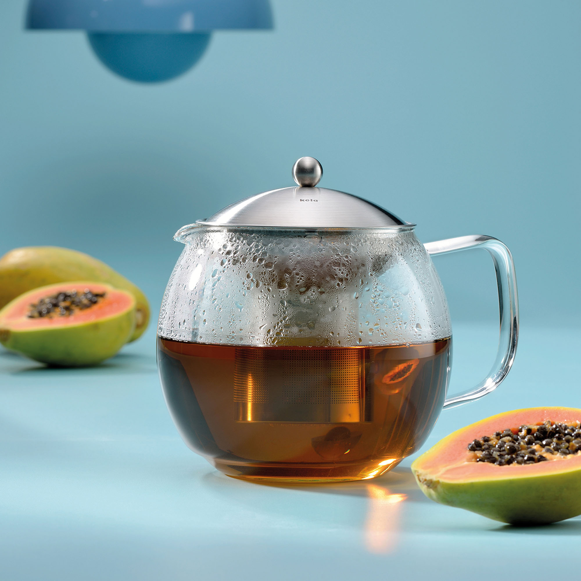 Transparente Teekanne mit bernsteinfarbenem Tee zwischen zwei aufgeschnittenen Papayas auf blauem Untergrund