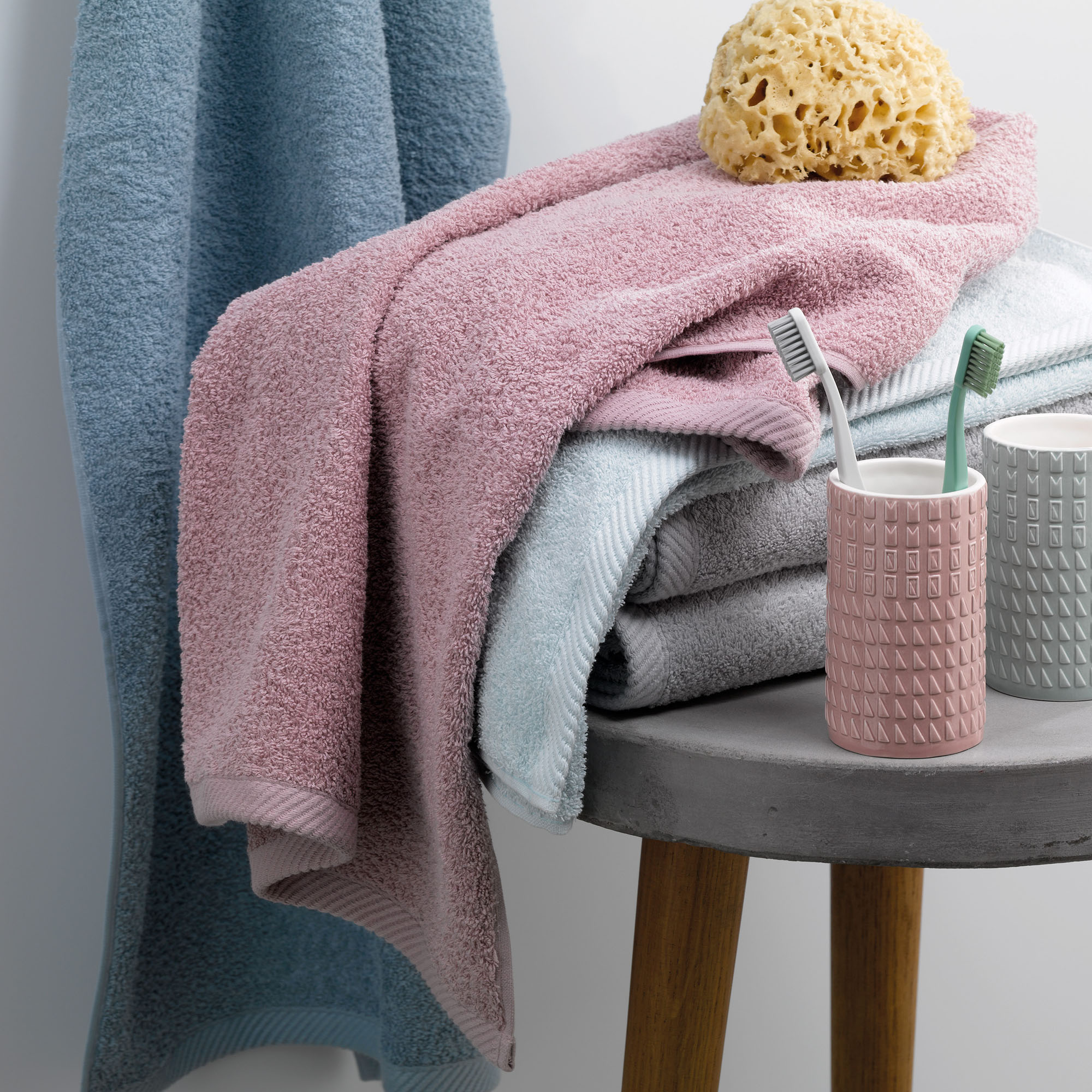 Altrosafarbenes Badtextil auf einem kleinen Beistelltisch neben Behältern Bürste und gelbem Badeschwamm