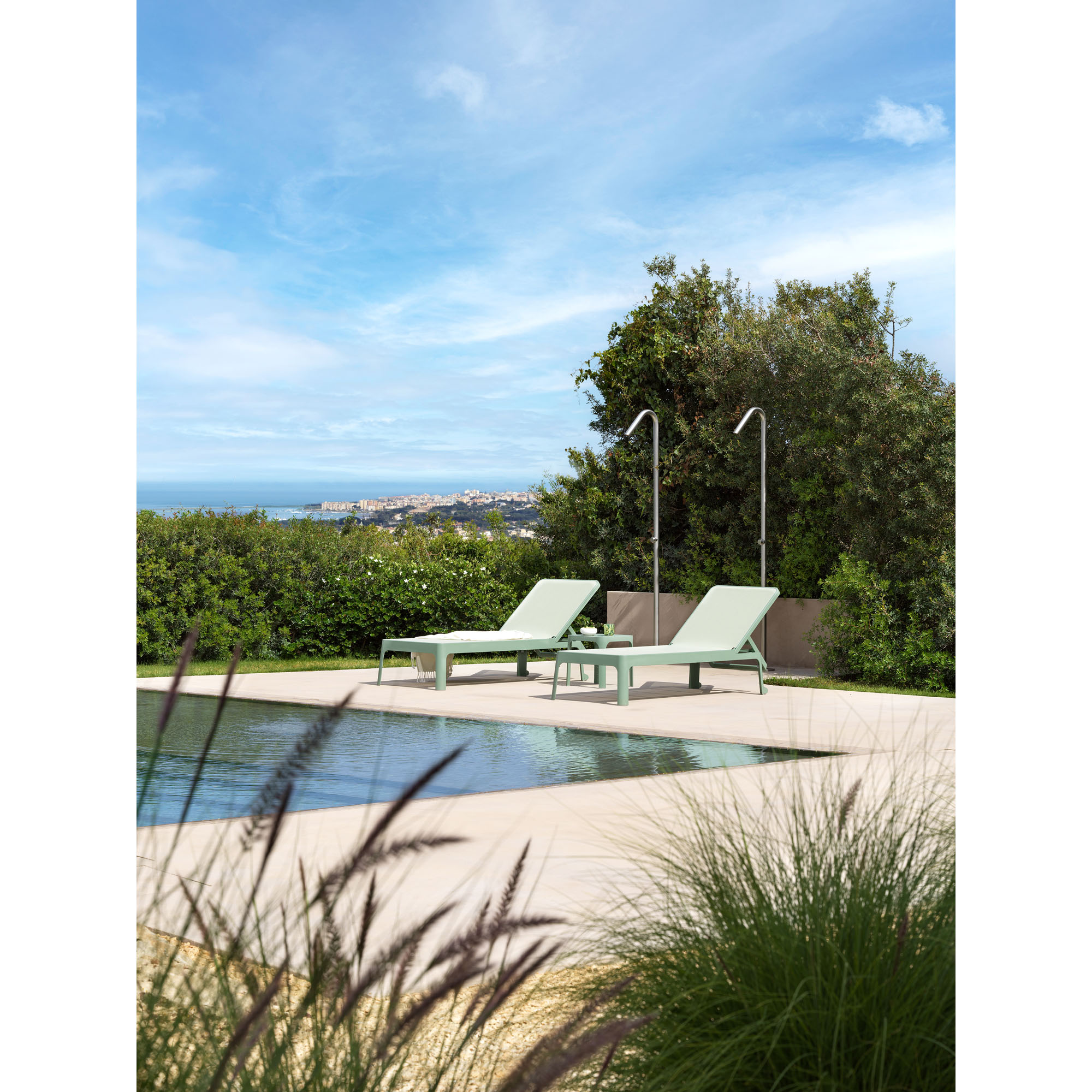 Zwei salbeigrüne Sonnenliegen stehen nebeneinander auf einer Terrasse am Pool. Im Hintergrund sind Büsche, Bäume und eine Aussicht auf eine Stadtlandschaft zu sehen.