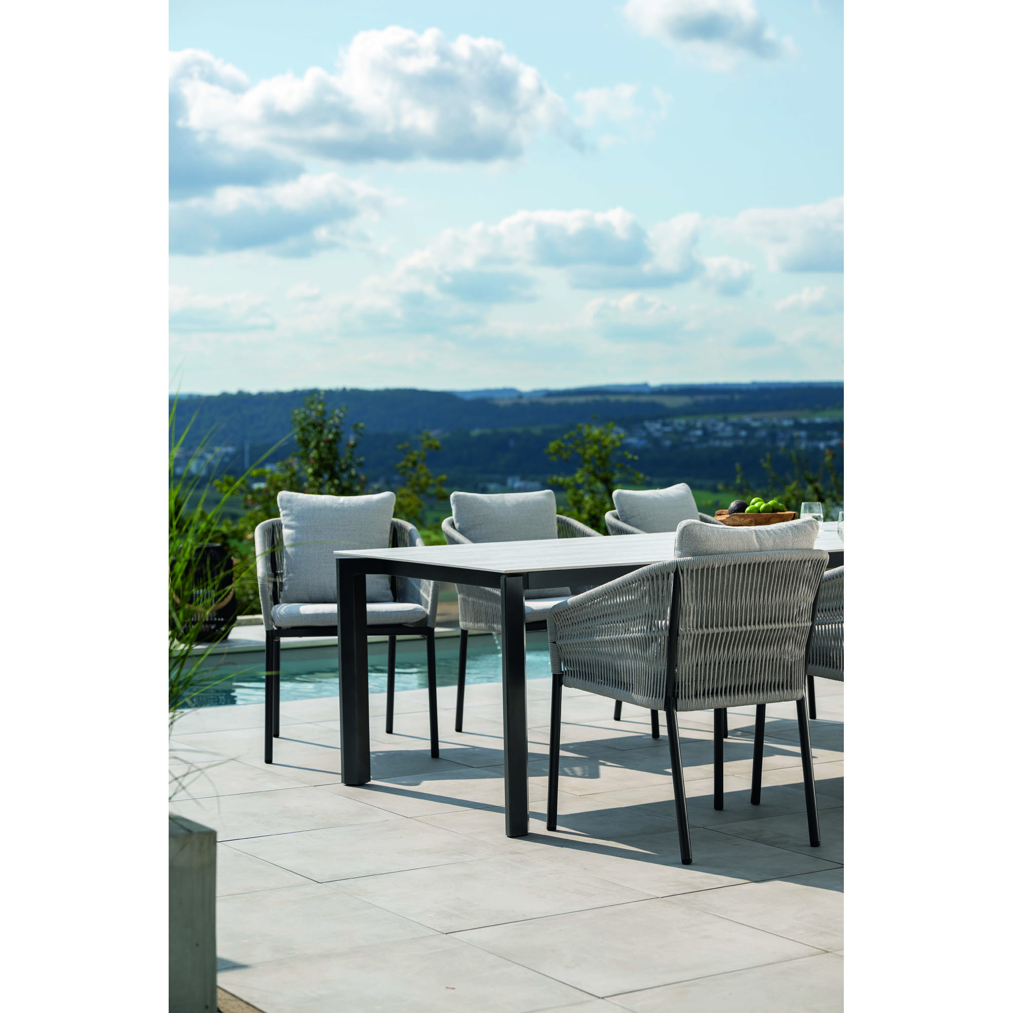 Ein Gartentisch mit mehreren Stühlen auf einer Terrasse, aus seitlicher Ansicht fotografiert, mit Blick ins Tal und bewölktem Himmel.