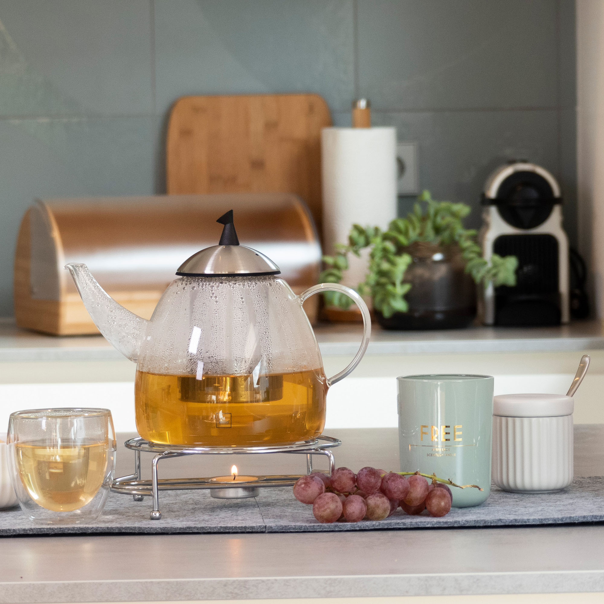 Transparente Teekanne mit Tee auf einem Tisch, daneben Tassen, Glas und Küchenutensilien