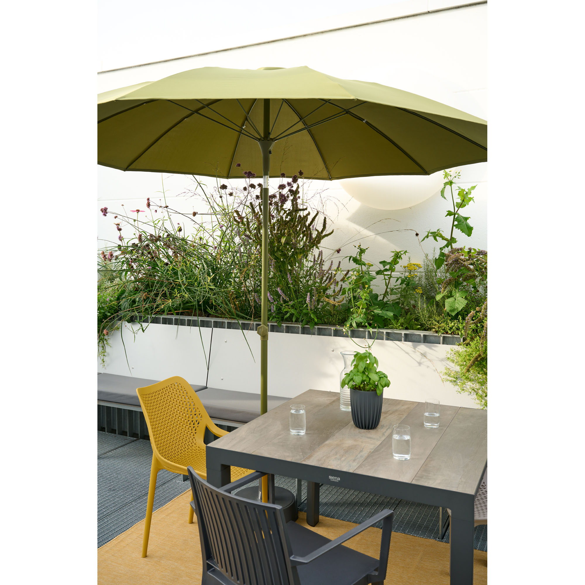Ein quadratischer Tisch mit Stühlen steht auf einer Terrasse unter einem grünen Sonnenschirm, im Hintergrund wachsen Pflanzen in Hochbeeten.