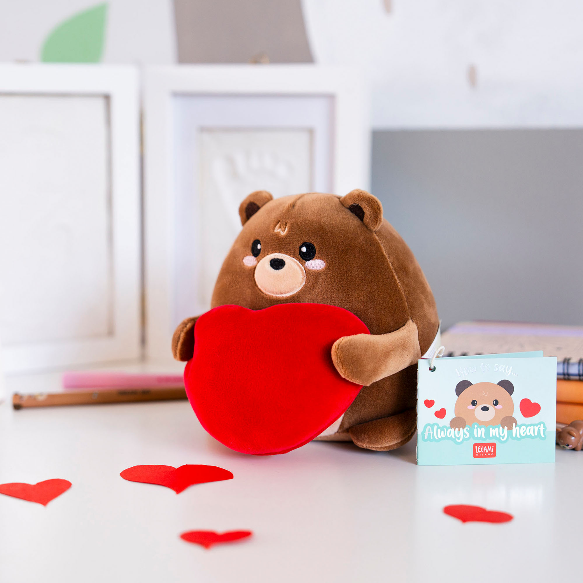 Braunes Teddybär-Plüschtier mit rotem Herz, auf einem Tisch zwischen roten Papierherzen dargestellt.