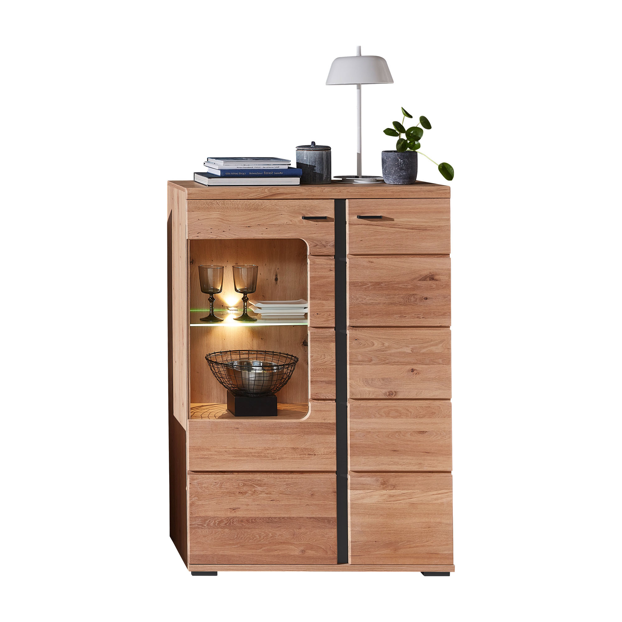 Frontansicht eines Sideboards in Holzoptik. Links befindet sich ein beleuchteter Glasausschnitt mit zwei Gläsern, Büchern und einer Metallschale. Auf der Oberseite stehen Bücher, eine Tischlampe und eine kleine Pflanze. Rechts sind geschlossene Türen sich