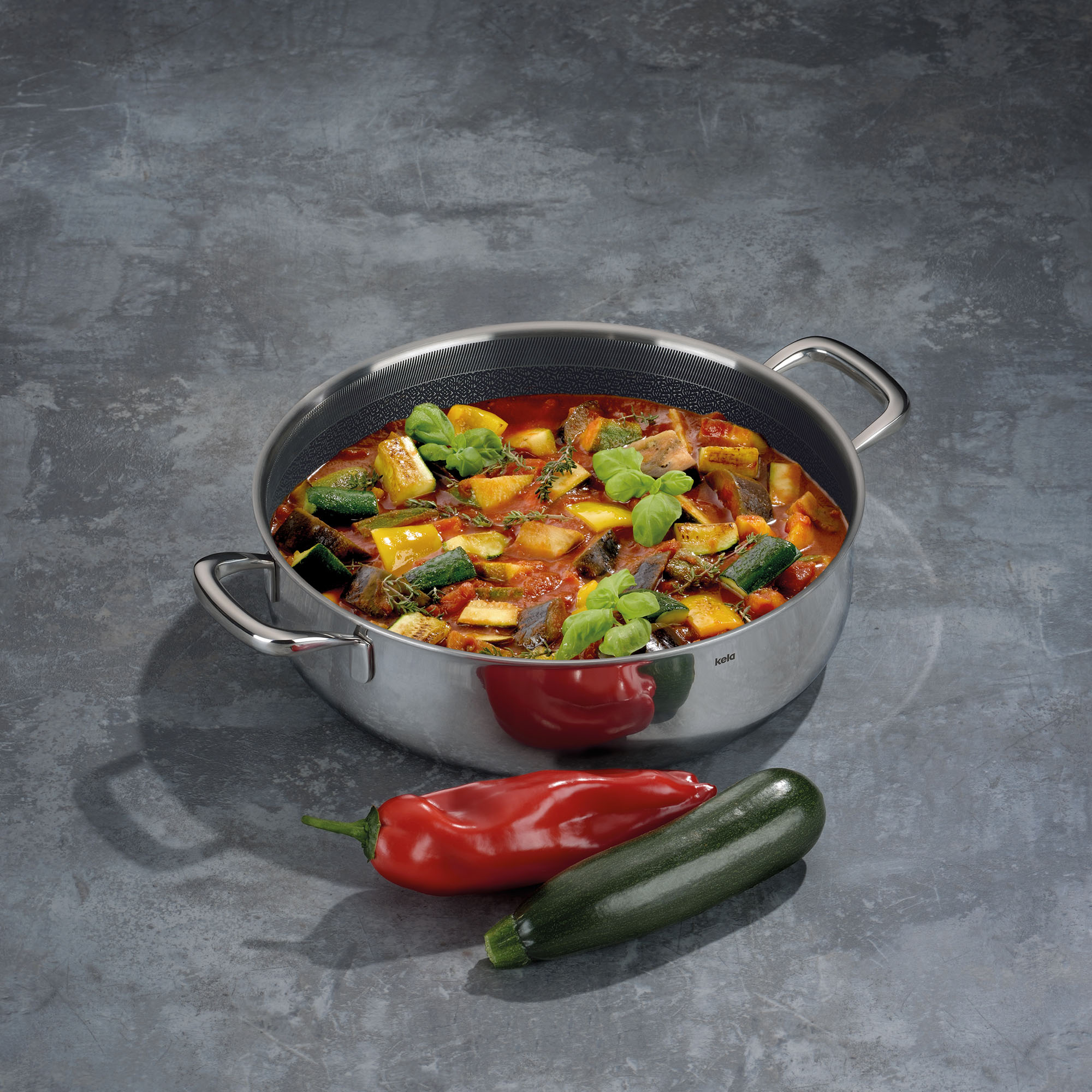 Dunkel beschichtete Servierpfanne mit buntem Gemüse auf einer grauen Arbeitsfläche neben Paprika und Chilischoten