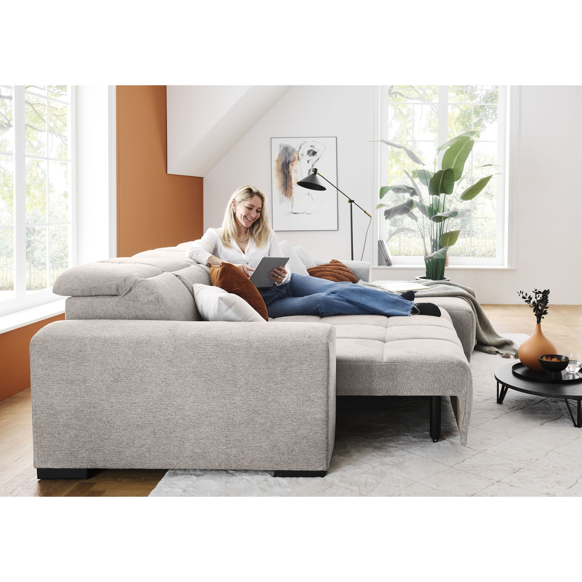 Frau liegt mit einem Tablet auf einem silbergrauen Schlafsofa mit ausgezogener Liegefläche, erhöhten Kopfstützen und mehreren Kissen im Wohnzimmer