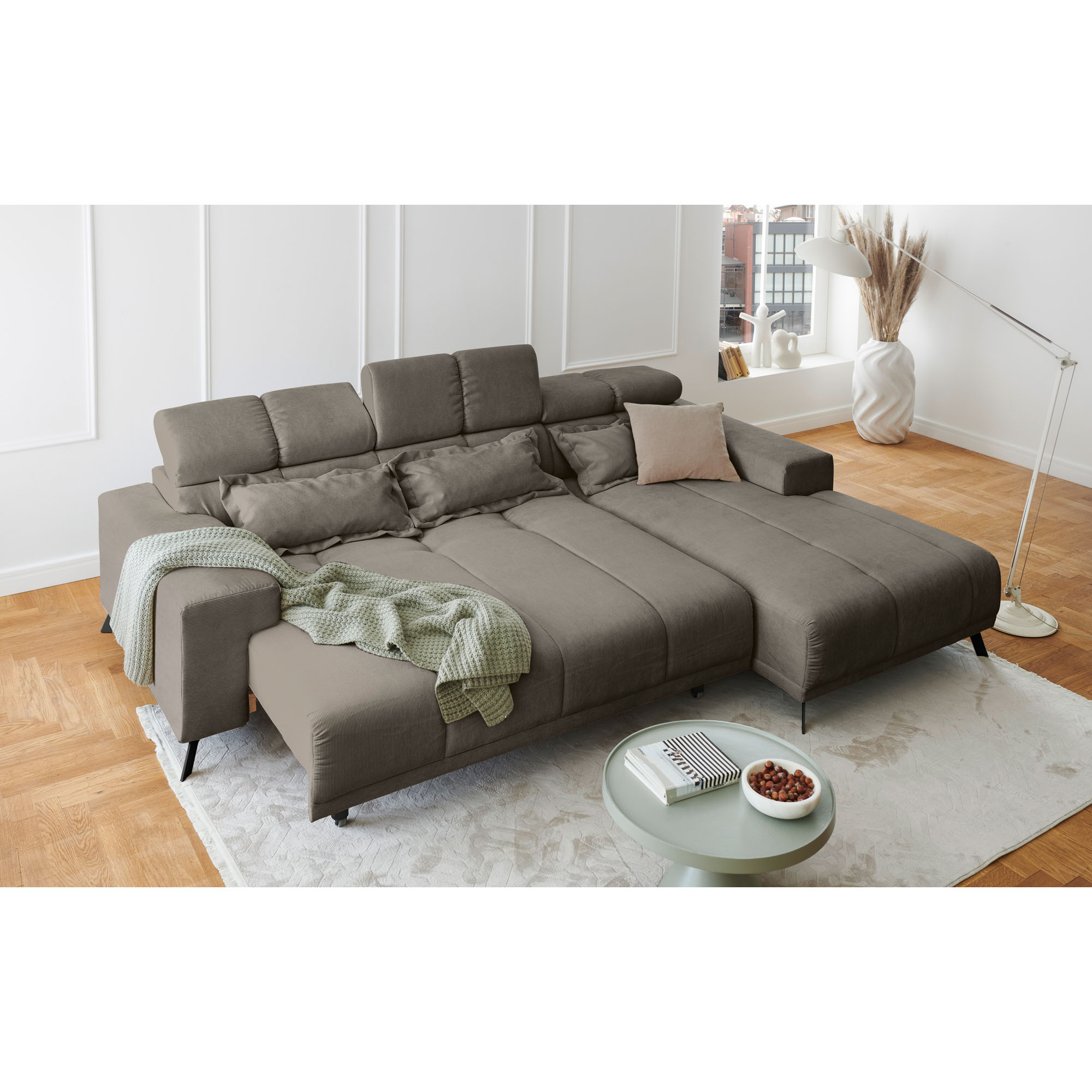 Hellgraues Ecksofa mit Ottomane rechts und ausgezogener Liegefläche in einem hellen Wohnzimmer mit Teppich und Couchtisch