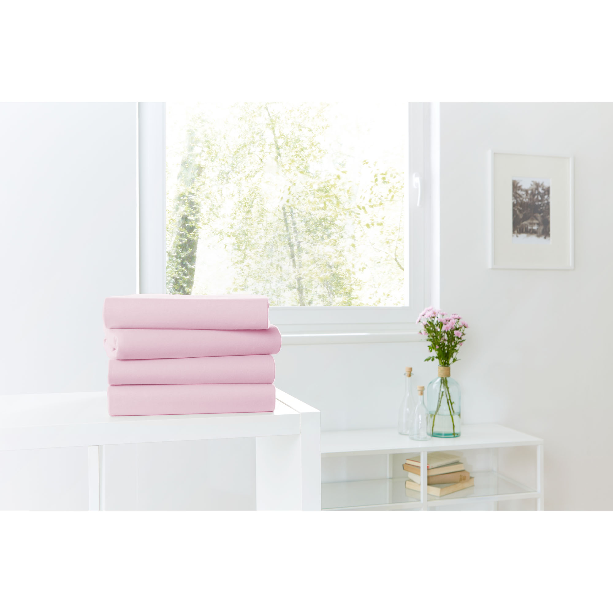 Vier gefaltete Spannbetttücher in Rosé auf einer weißen Kommode vor einem hellen Fenster; Größe 100 x 200 x 30 cm.
