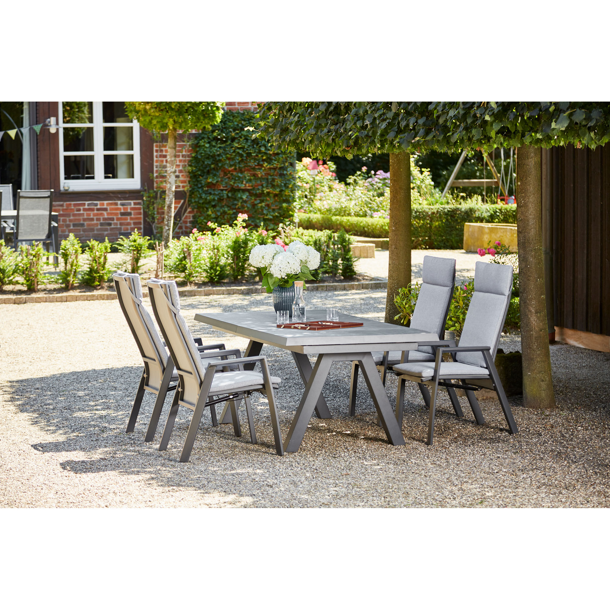 Mehrere Stühle mit grauer Polsterung stehen um einen großen rechteckigen Tisch im Garten, im Hintergrund Pflanzen, Bäume und ein Haus.