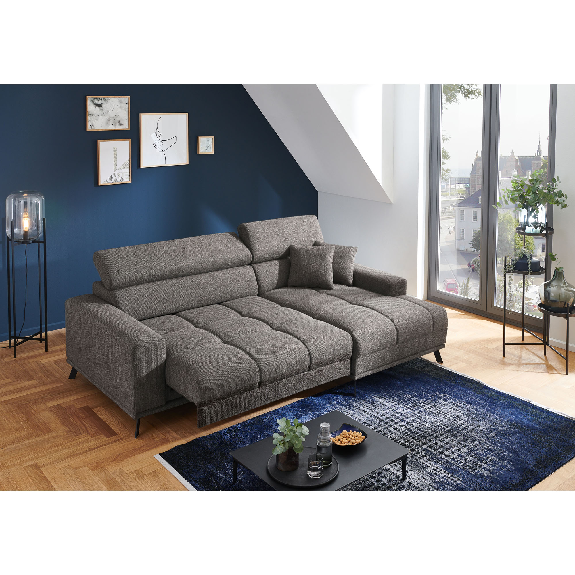 Dunkelgraue Wohnlandschaft mit Longchair rechts im Bild, vorgezogenen Sitzflächen und verstellbaren Kopfstützen in einem hellen Wohnraum