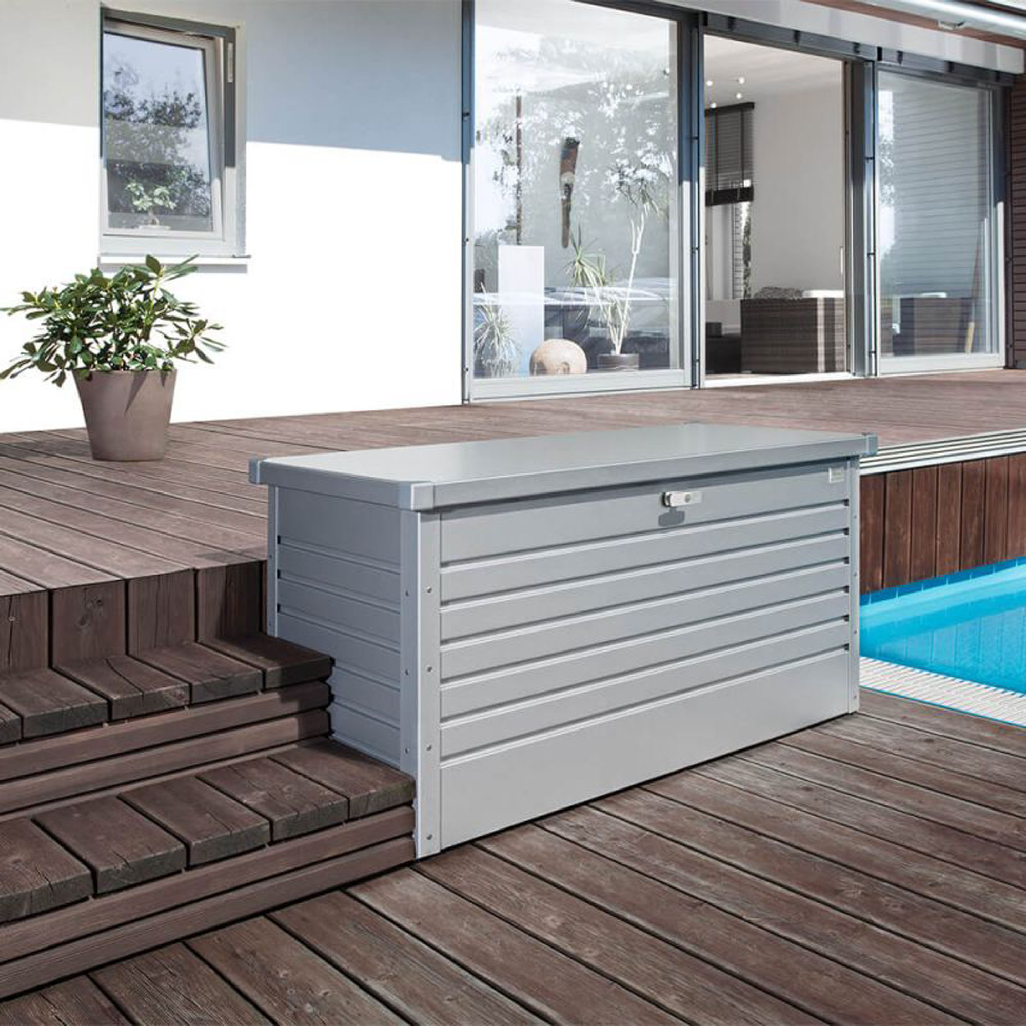 Silberfarbene Auflagenbox aus Metall auf einer Holzterrasse neben einem Pool vor einem Wohnhaus