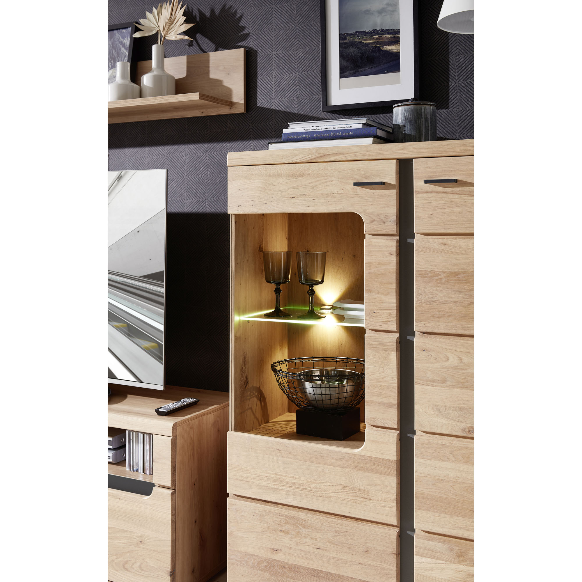 Detailaufnahme einer Vitrine in Holzoptik. Zu sehen sind zwei beleuchtete Glasböden mit Gläsern und einer dekorativen Metallschale. Im Hintergrund hängt ein Wandboard mit Vasen und einem Bild.