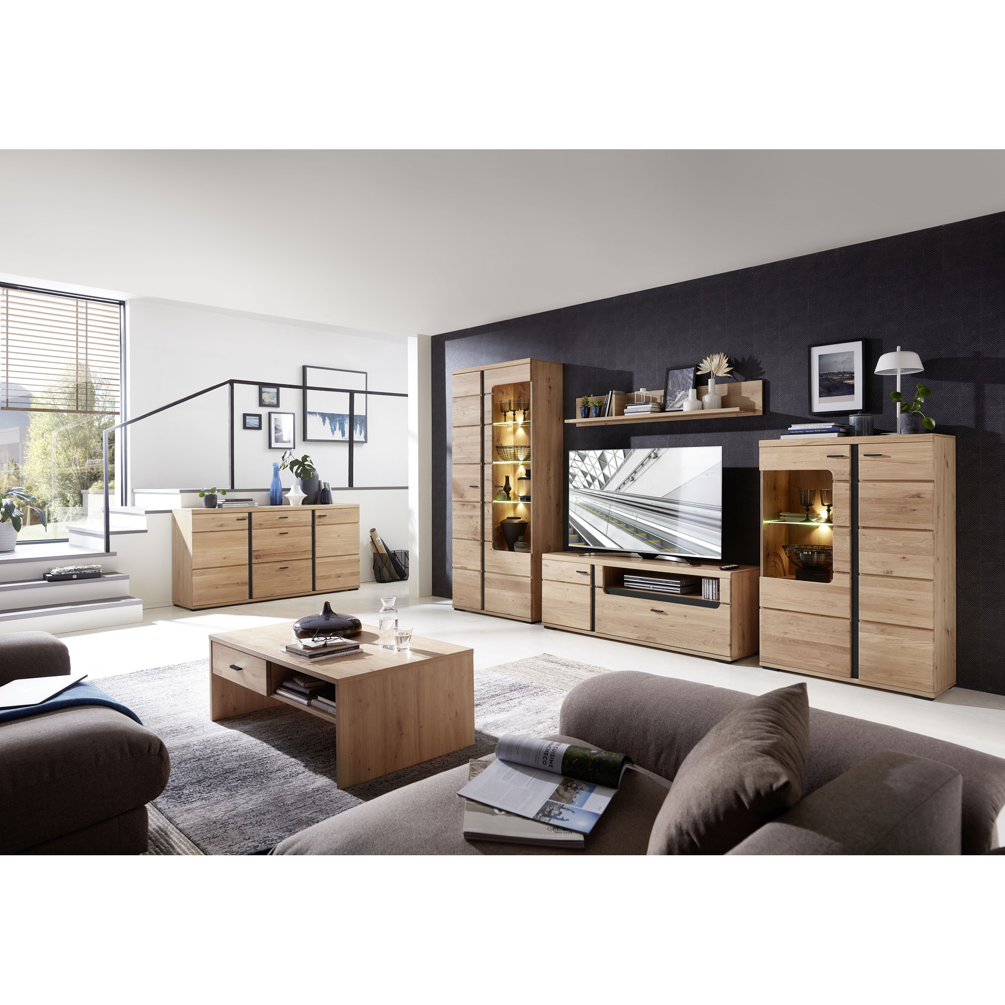 Großes Wohnzimmer mit Couchtisch aus hellem Holz, Sofas, Teppich und einer Wohnwand; Kommode, Vitrine und Fernseher sind sichtbar, Tageslicht fällt durch ein großes Fenster ein.