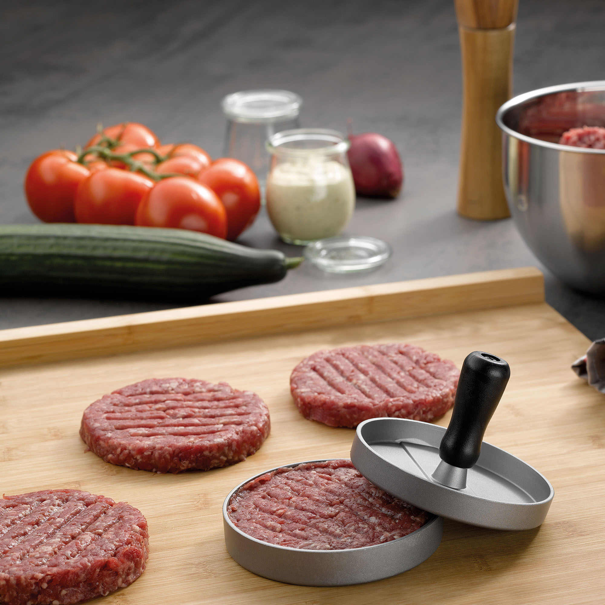 Bambus Schneidebrett mit Burger Patties Tomaten und Küchenutensilien auf einer Arbeitsplatte