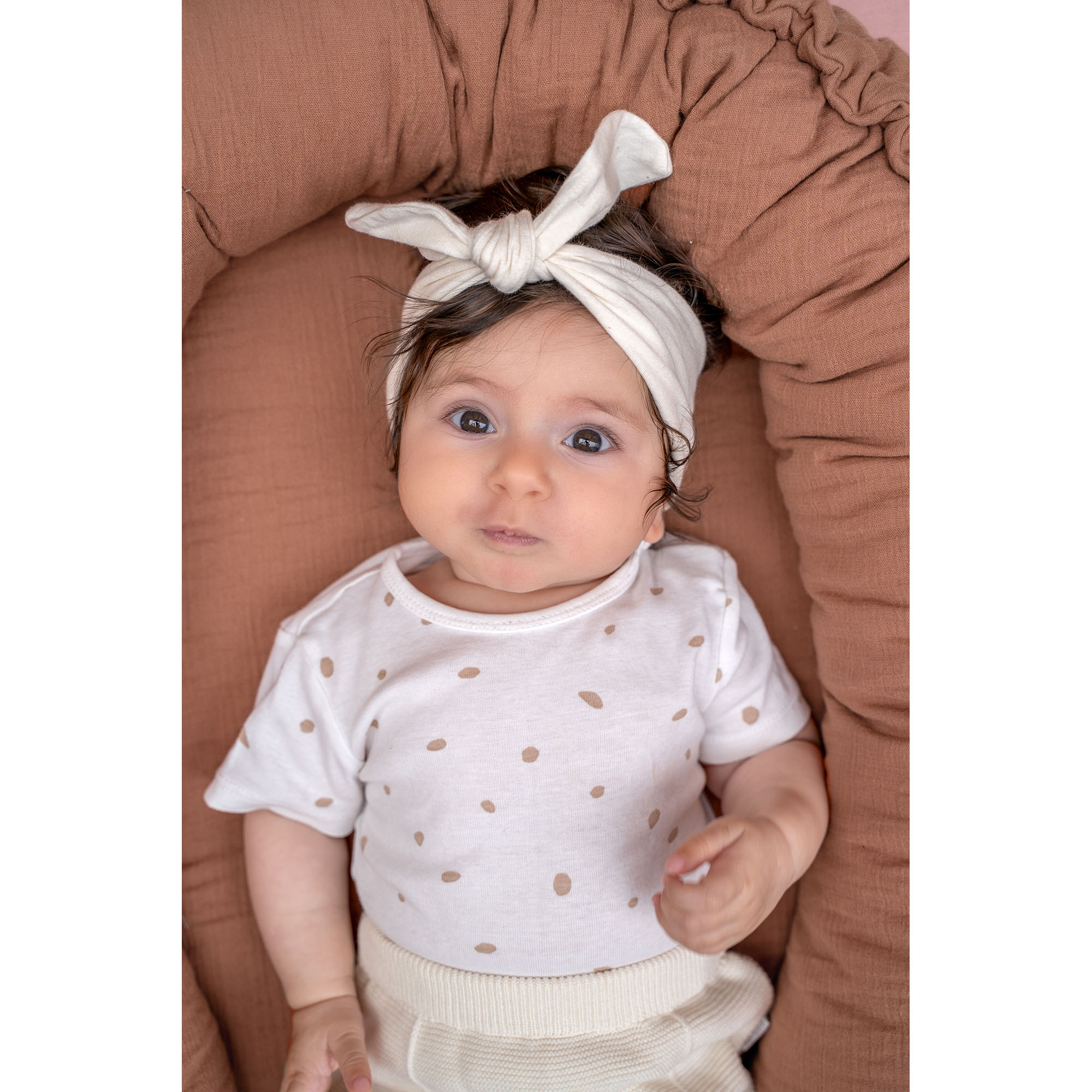 Baby liegt im rostbraunen Babynest aus Musselin, trägt ein cremefarbenes Shirt mit Punkten und ein Stirnband mit Knoten