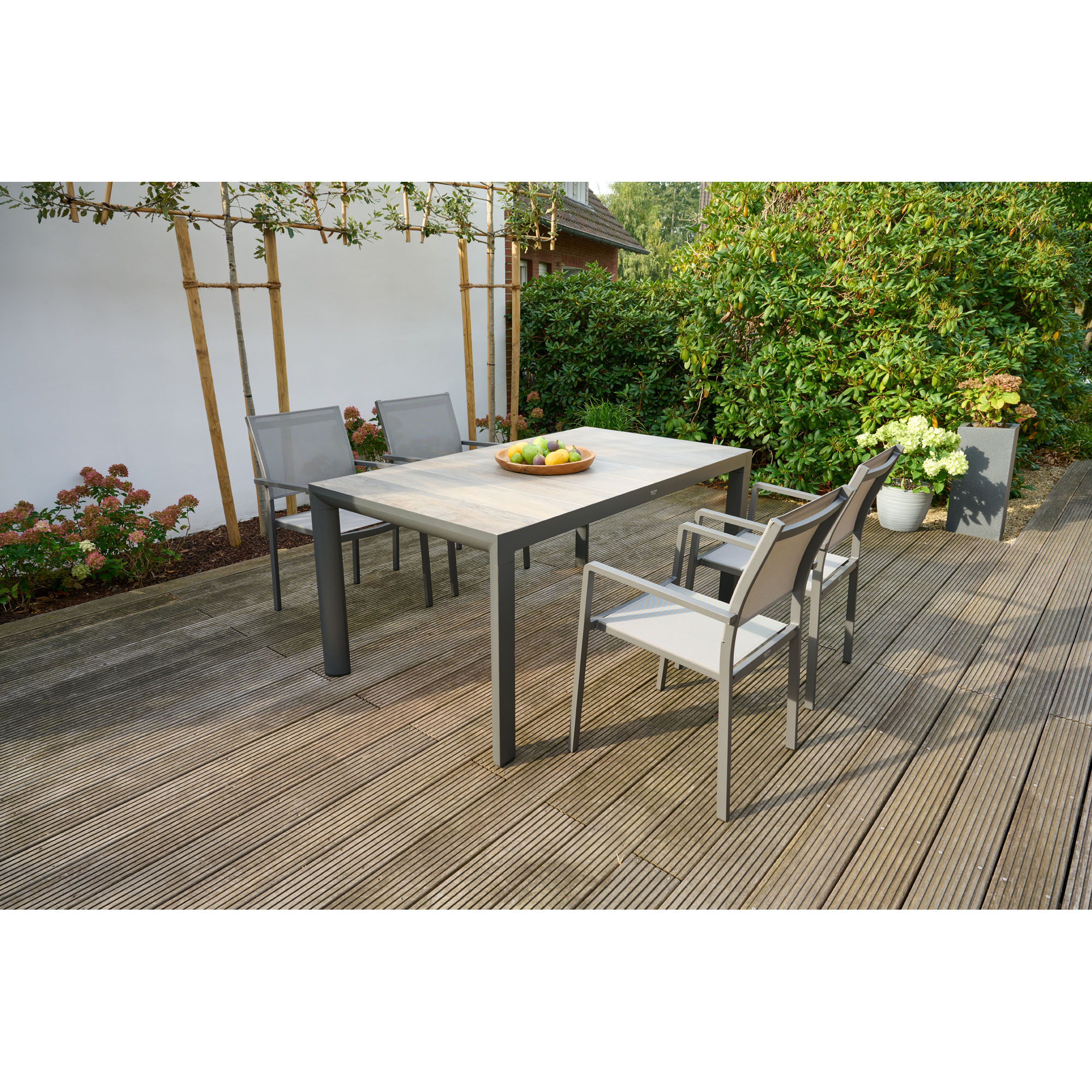 Gartentisch mit Holzdekor-Platte und mehreren Stühlen auf einer Holzterrasse im Garten, von Pflanzen umgeben.