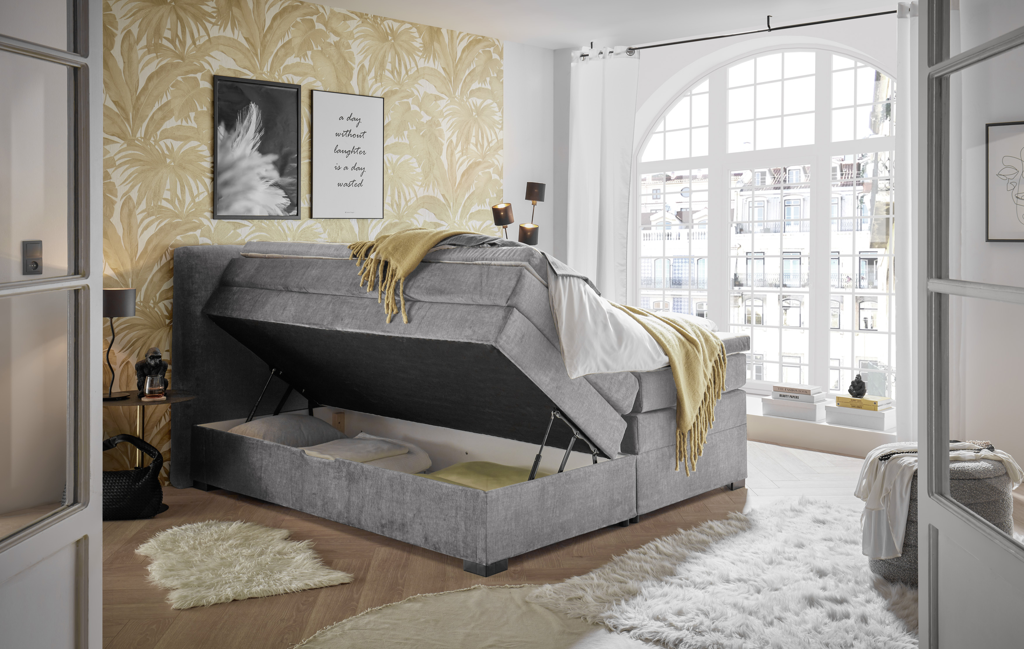 Boxspringbett „Vogue 15 grey“ in einem Schlafzimmerambiente, Bettkasten geöffnet, dekoriert mit Kissen und Bettdecke
