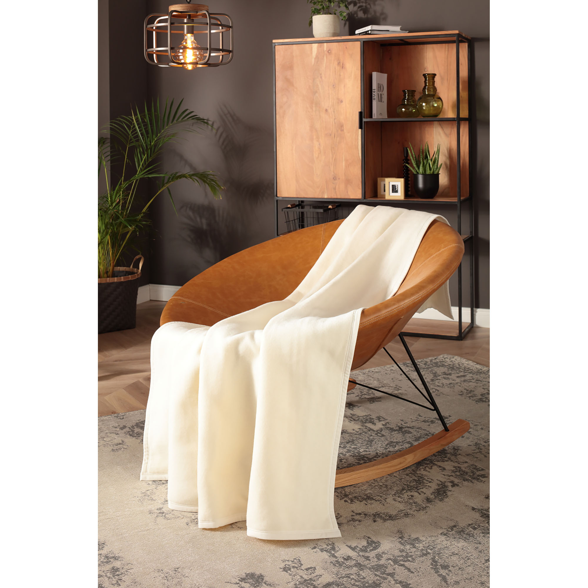 Wohndecke CottonSense natur fällt in langen cremefarbenen Falten über einen cognacfarbenen Loungesessel.