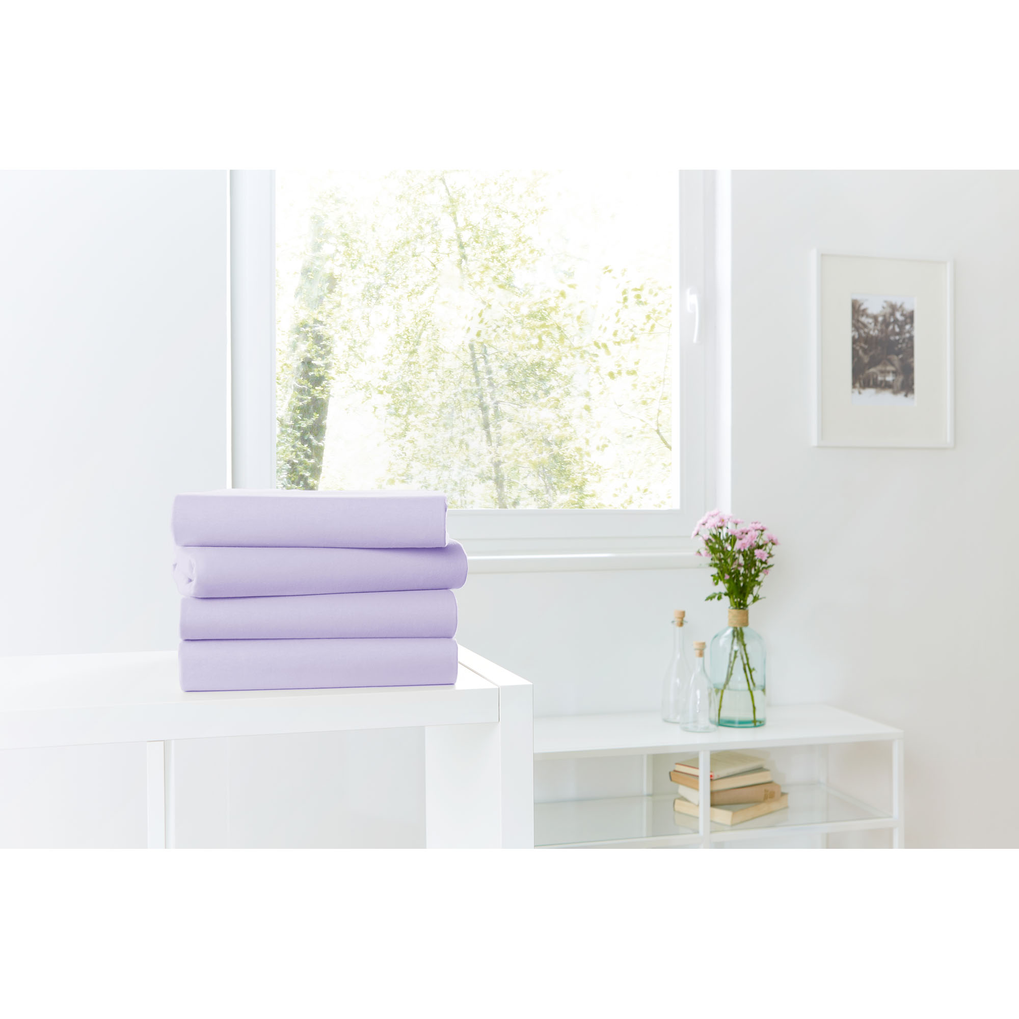 Vier gefaltete Spannbetttücher in Lavendel auf einer weißen Kommode vor einem hellen Fenster; Größe 100 x 200 x 30 cm.