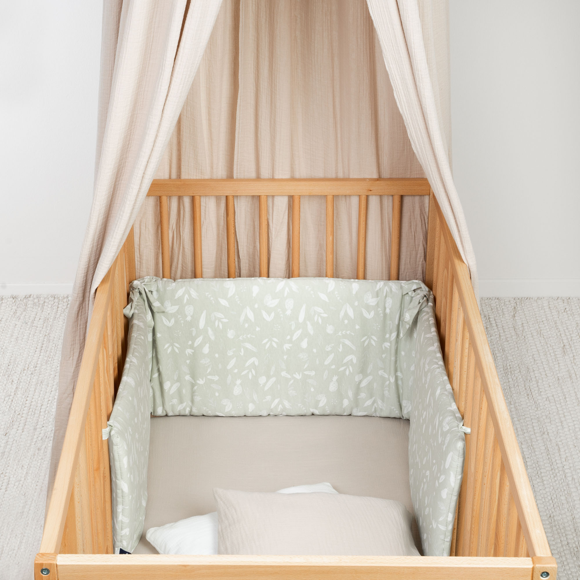 Holz-Babybett mit olivgrünem Nestchen mit weißem Blättermuster und beigem Baldachin, innen mit Kissen und Decke ausgestattet