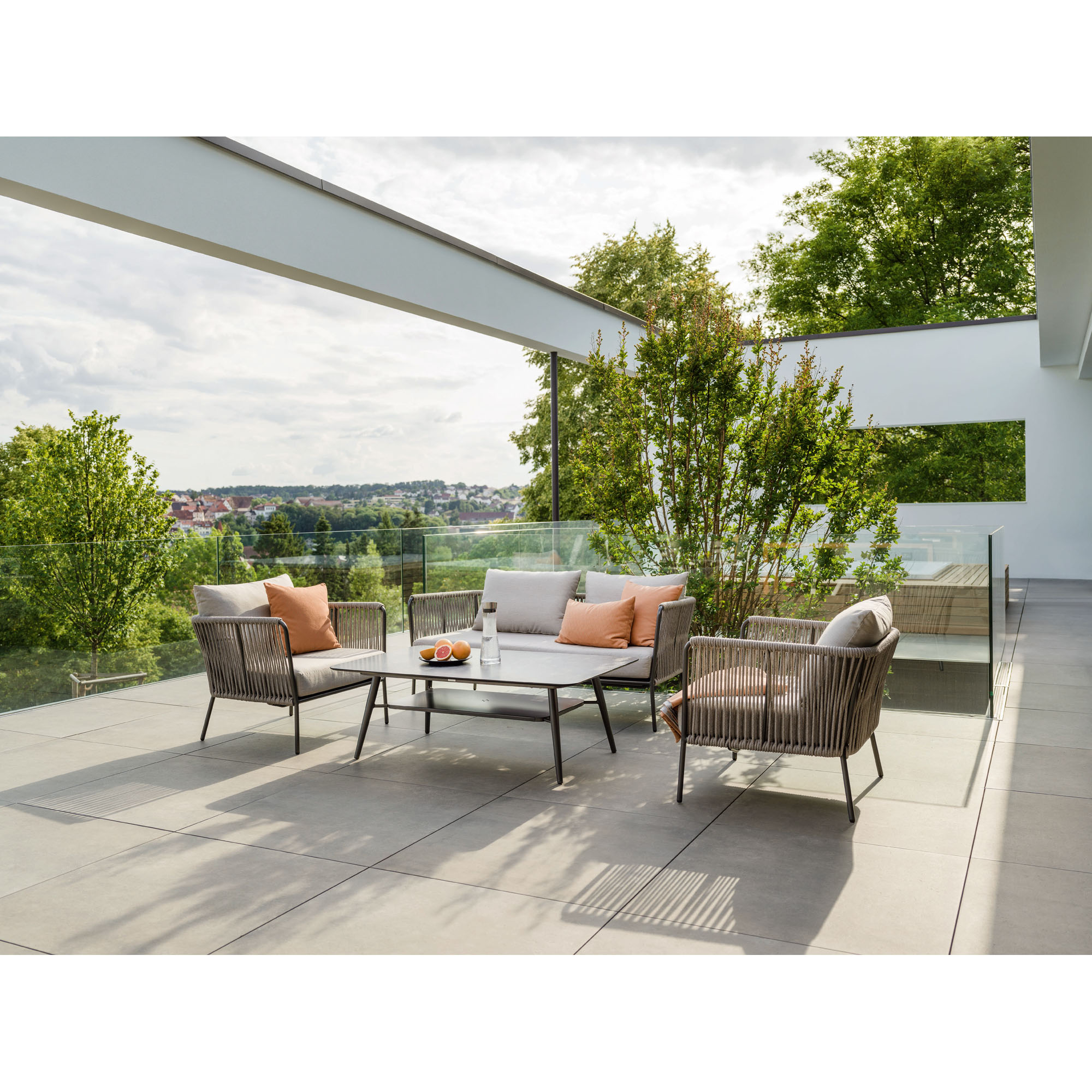 Eine Terrasse mit einem Gartenset aus Sofa, Sesseln und Tisch. Die Möbel haben helle Polster und Seilbespannung. Im Hintergrund sind Glasgeländer, Bäume und eine weite Aussicht auf Häuser und Landschaft zu sehen.