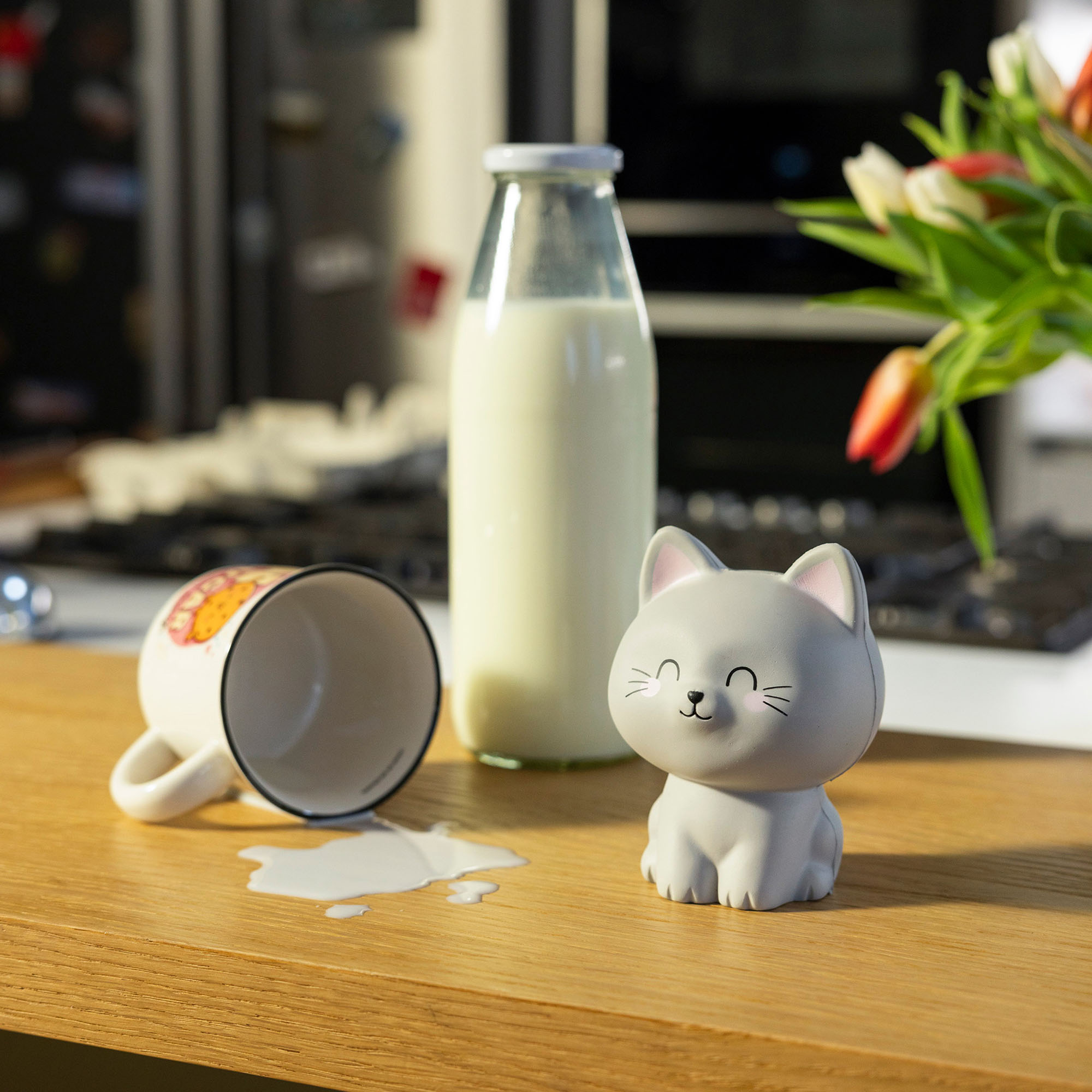 Graue Antistress-Figur in Katzenform, auf einem Holztisch neben Flasche und umgekipptem Becher gezeigt.