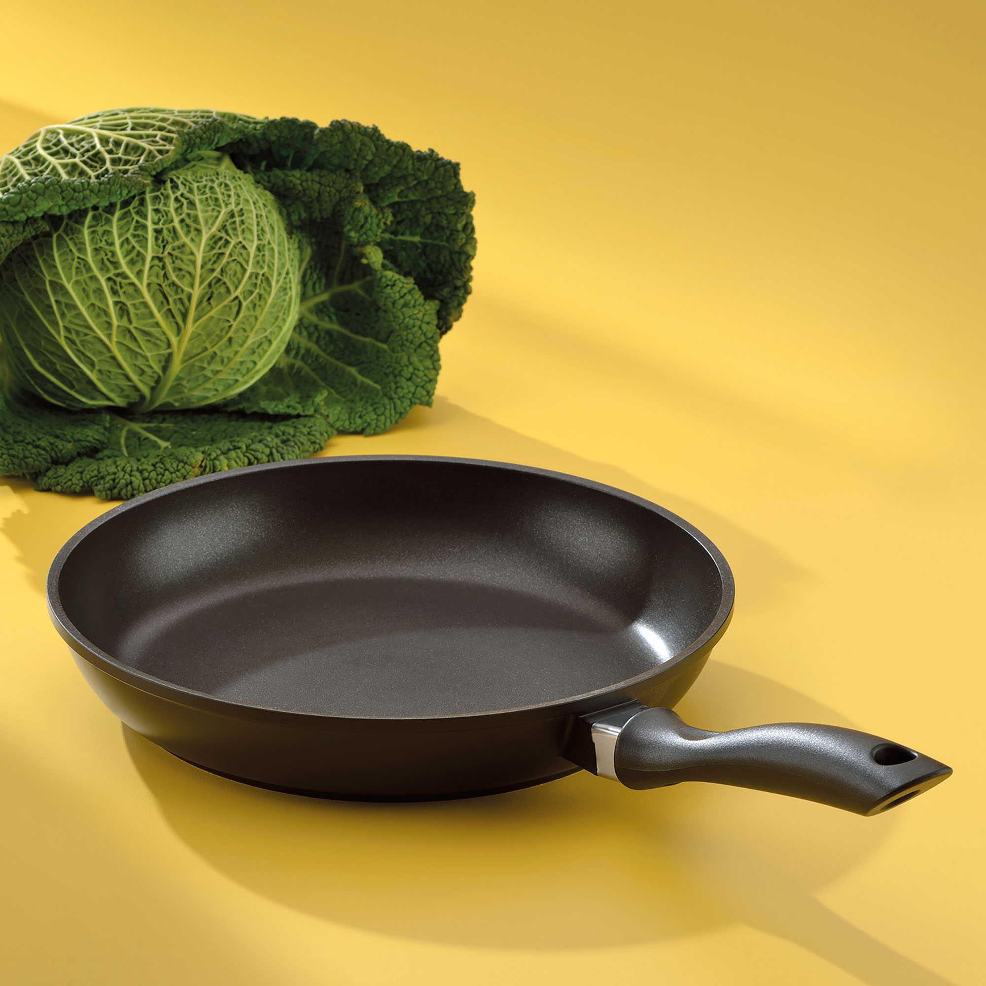 Leere schwarze Bratpfanne mit langem Griff neben einem grünen Wirsing auf gelbem Untergrund