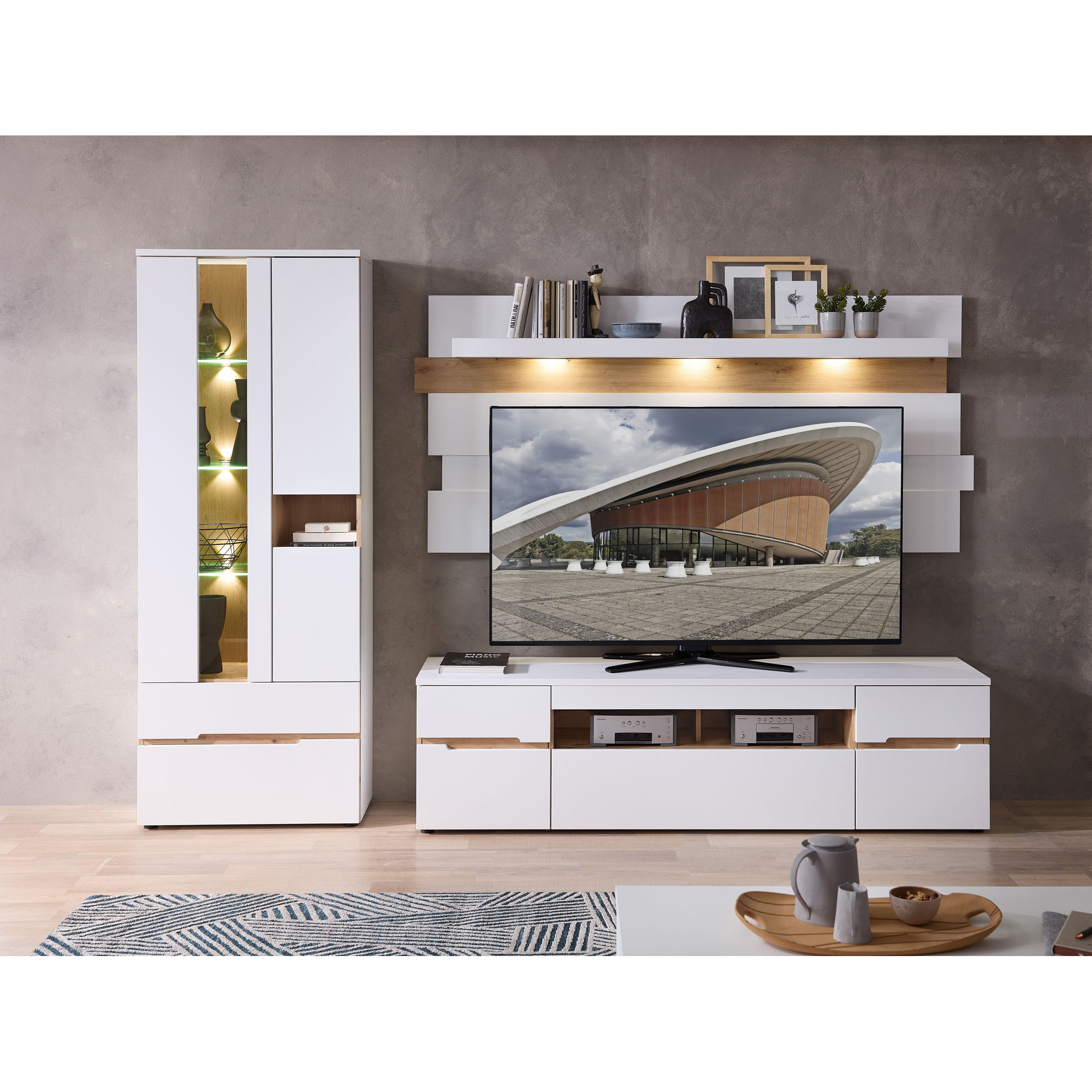 Wohnzimmeransicht mit einer weißen Wohnwand. Hinter dem Fernseher befinden sich weiße Wandpaneele und darüber ein beleuchtetes Wandboard in Holzoptik. Links steht eine Vitrine mit beleuchteten Glasfächern und geschlossenen Elementen. Auf dem Wandboard ste