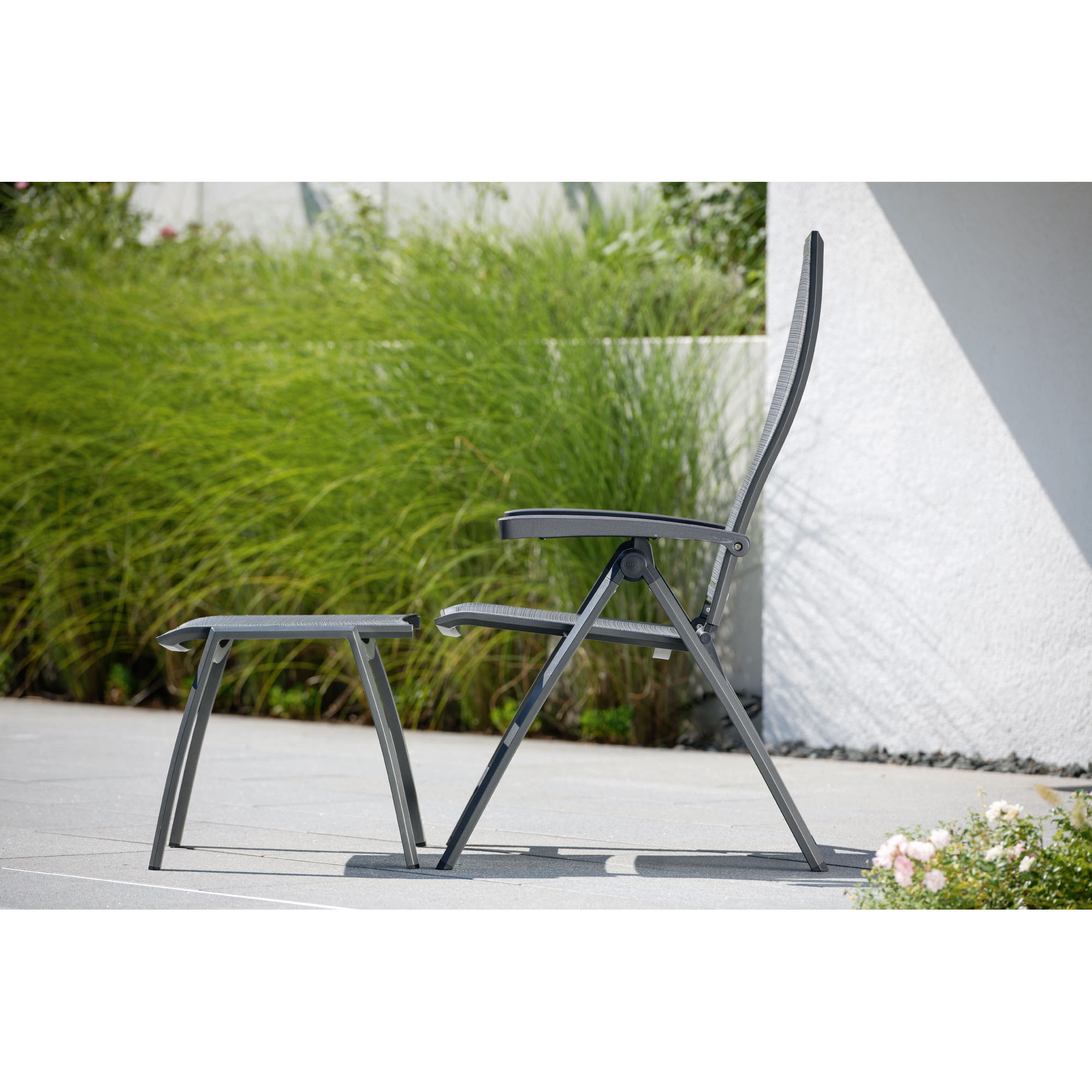 Ein seitlich fotografierter Gartenstuhl mit Hocker aus grauem Metall und Textilbespannung. Im Hintergrund sind hohe Gräser und ein heller Hausputz zu sehen.