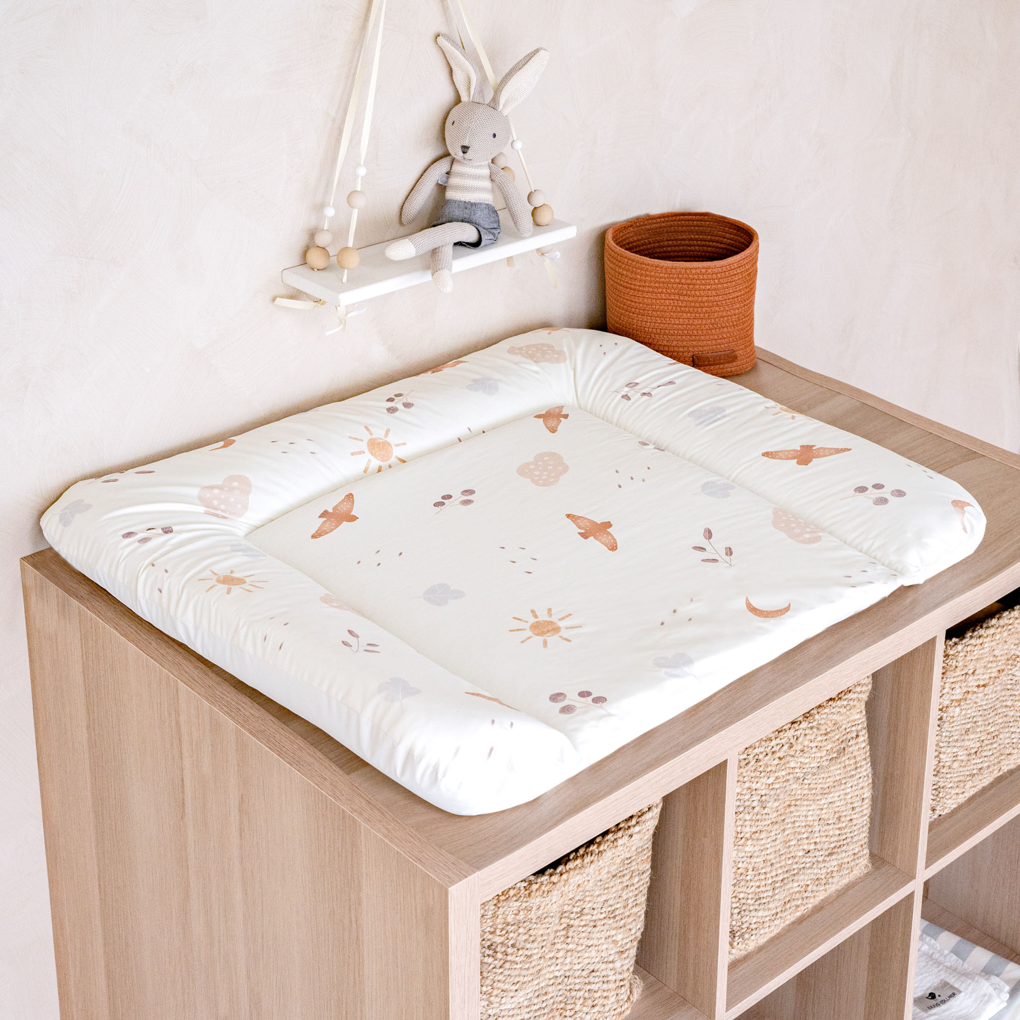 Beige Wickelauflage mit Vogel- und Sonnenmuster auf einer Holzkommode, daneben Korb und Stoffhase auf Wandregal