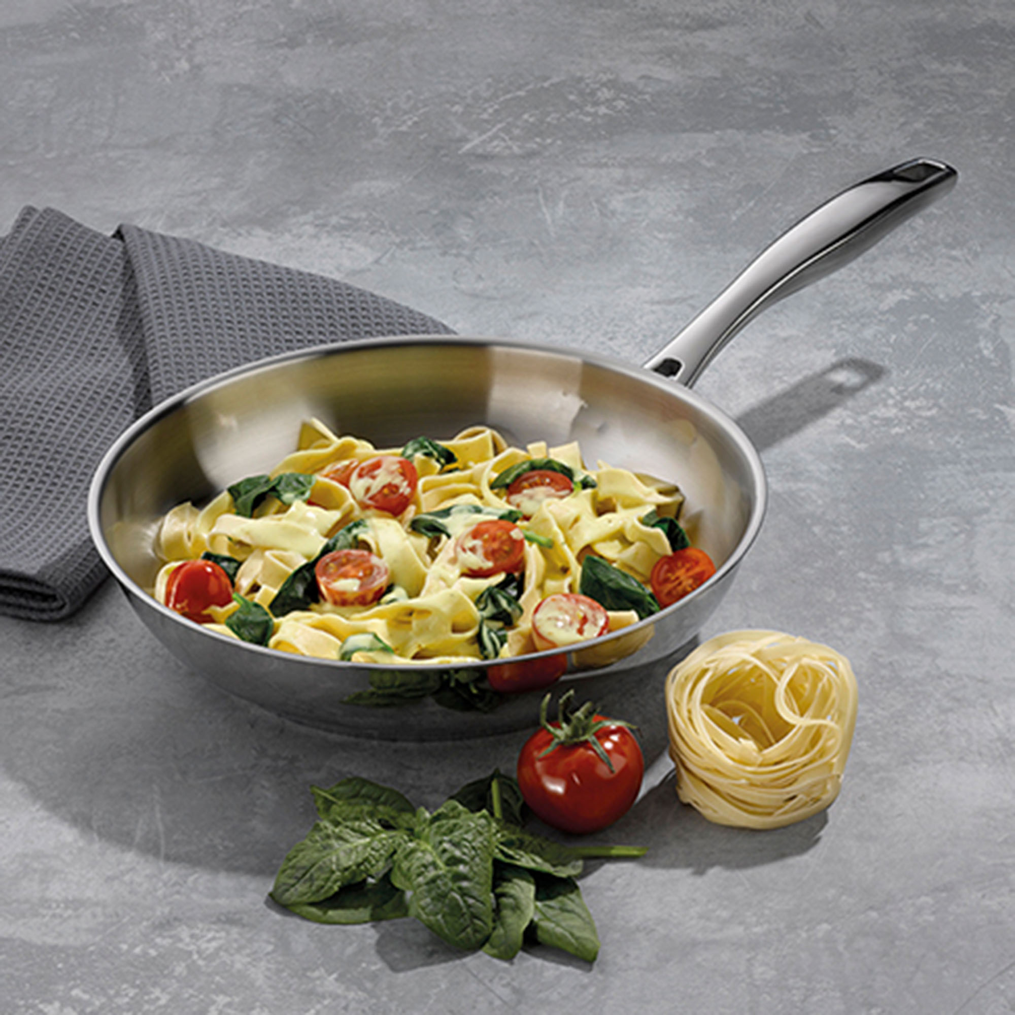 Edelstahl-Bratpfanne mit zubereitetem Gemüse, Kartoffeln und Tomaten auf einer grauen Arbeitsfläche