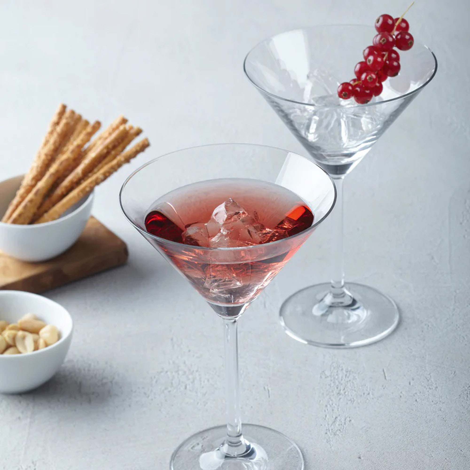 Martiniglas mit rotem Cocktail und Eiswürfeln daneben ein weiteres Glas mit roten Beeren sowie Snacks im Hintergrund