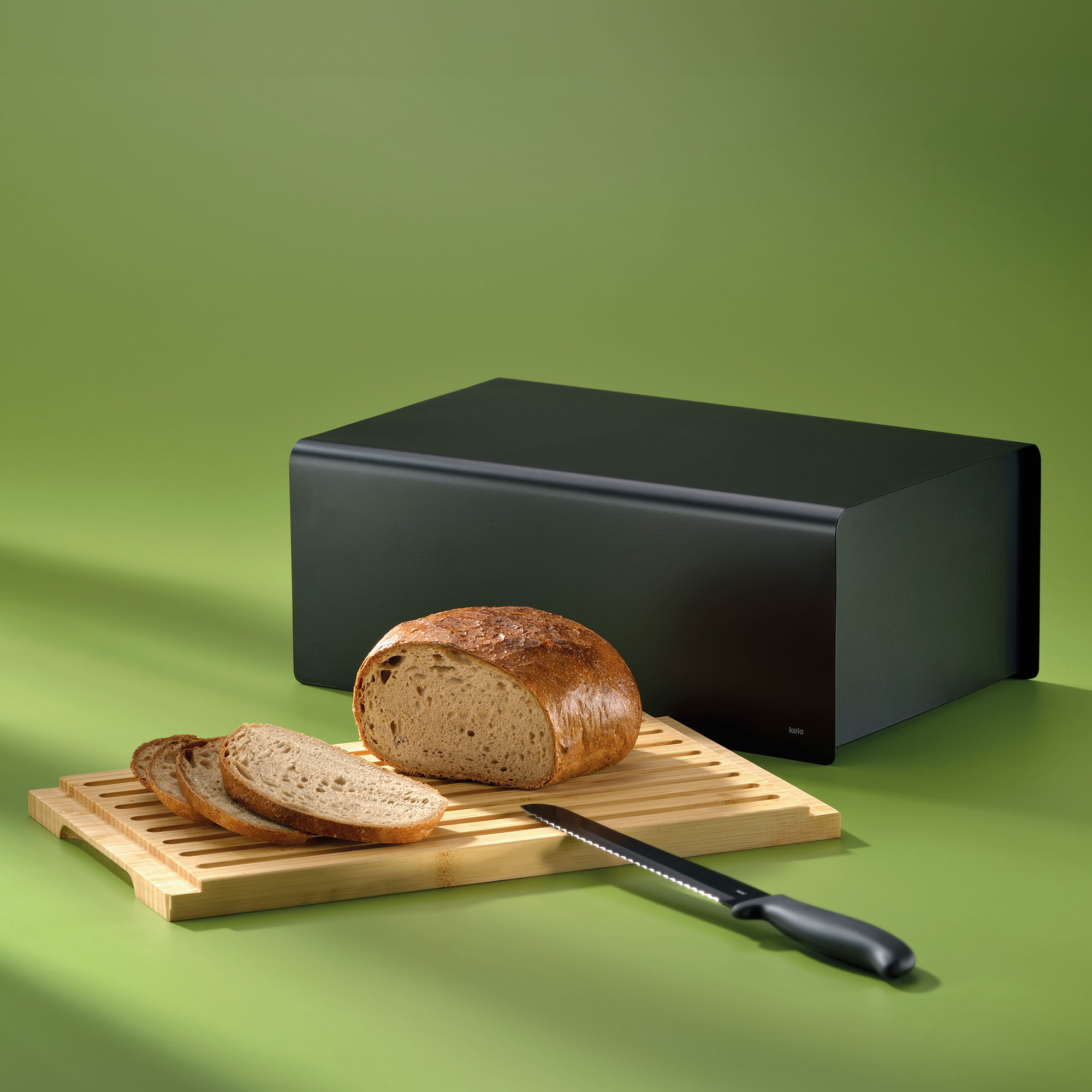 Schwarzer Brotkasten mit Brotlaib Messer und herausgezogenem Bambusbrett auf grünem Untergrund