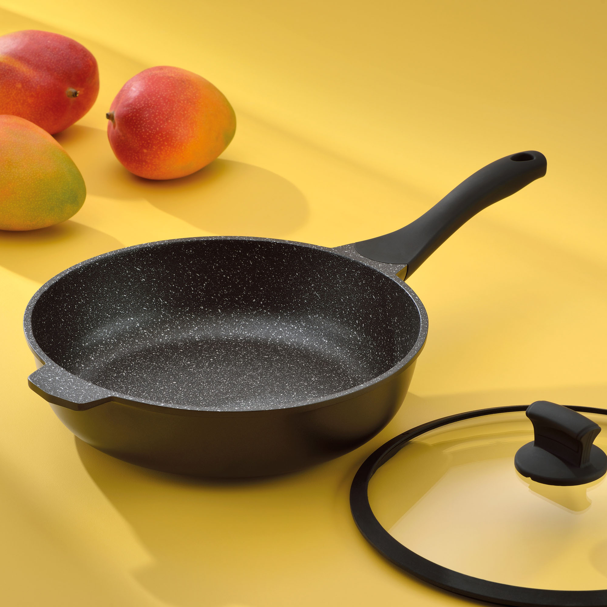 Schwarz beschichtete Bratpfanne mit langem Griff auf gelber Fläche neben einem Glasdeckel und roten sowie gelben Früchten