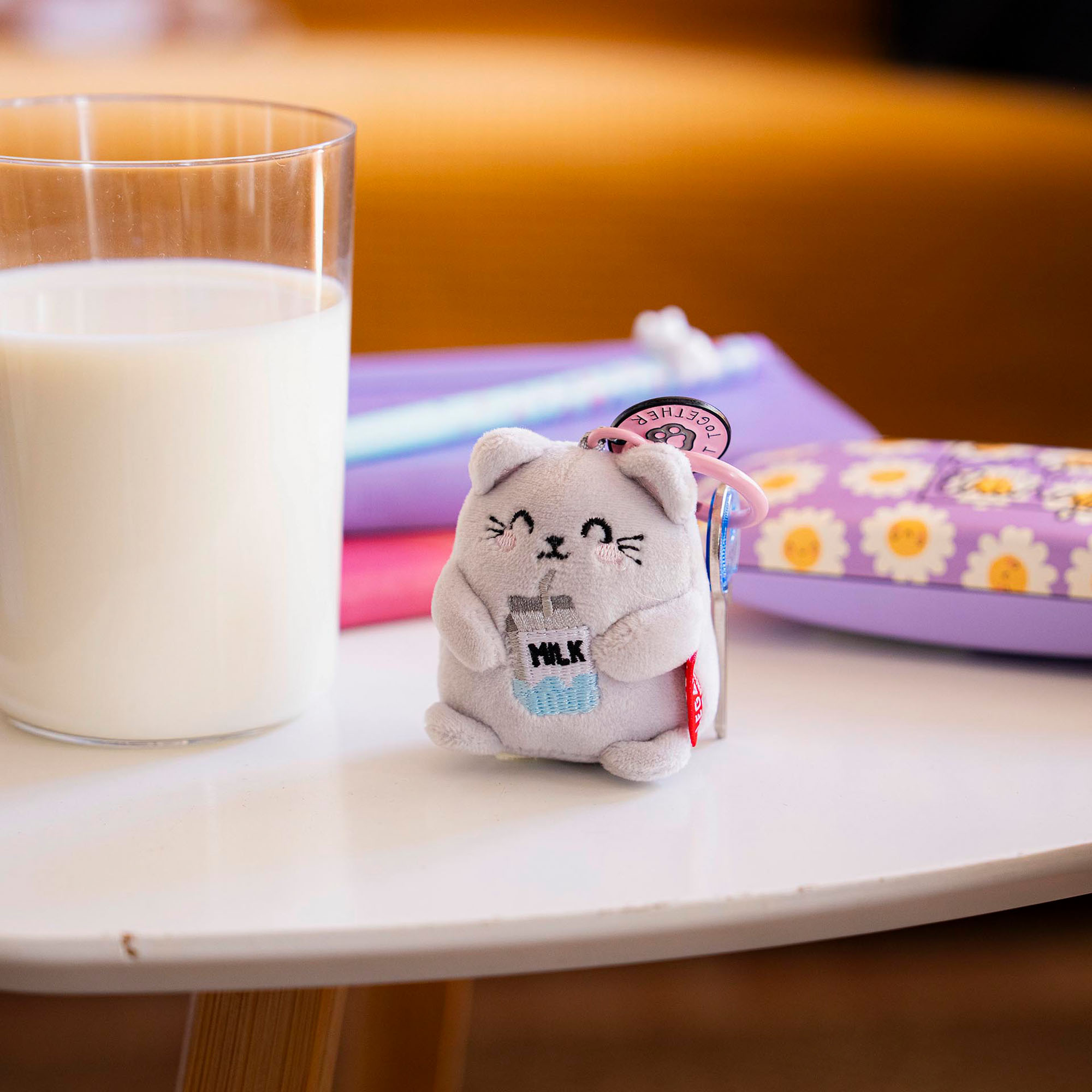 Grauer Katzen-Plüsch-Schlüsselanhänger mit Milchflasche, auf einem Tisch neben einem Glas Milch und bunten Stiften dargestellt.