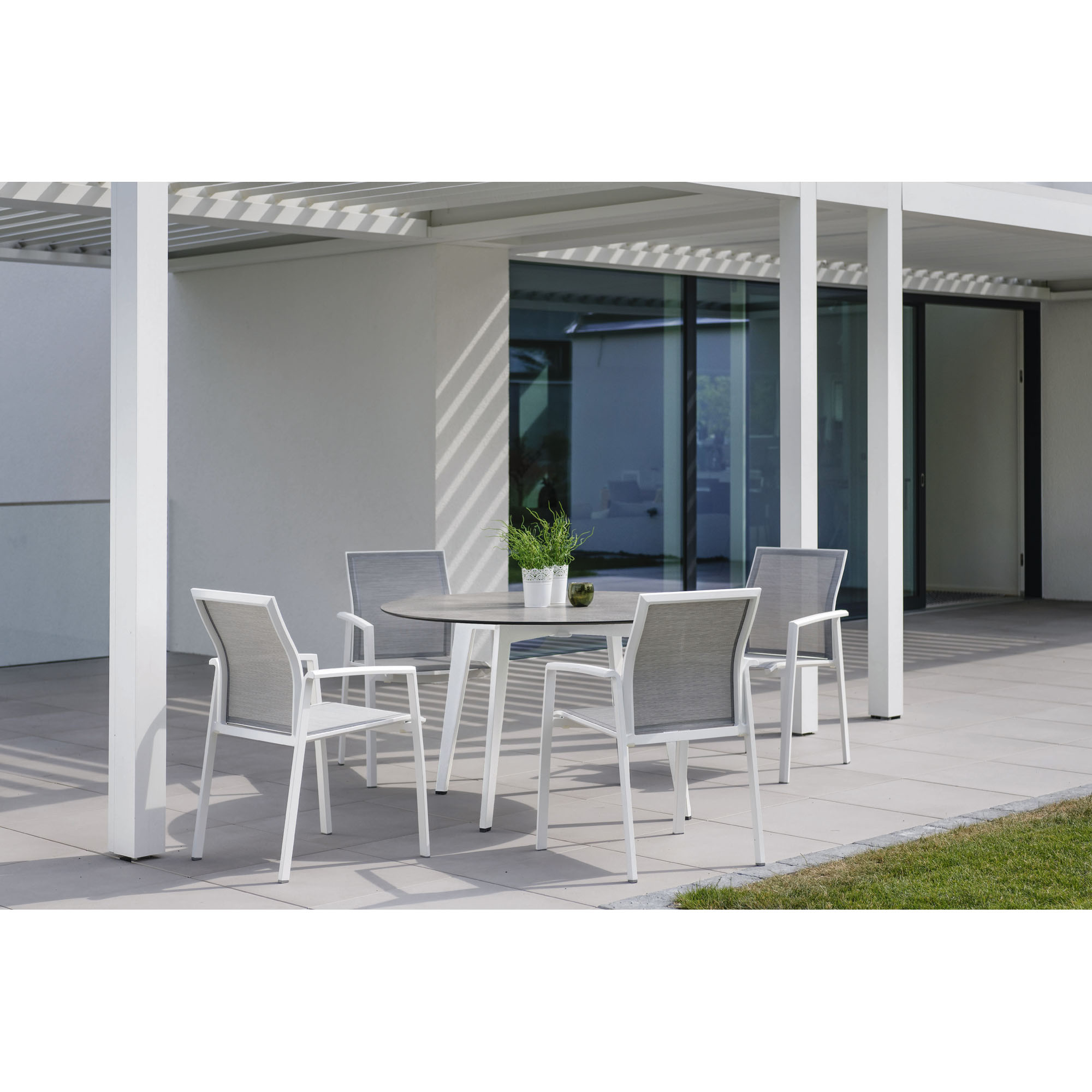 Eine Sitzgruppe mit vier weißen Gartenstühlen und einem runden Tisch steht auf einer hellen Terrasse vor einem modernen Gebäude mit Glasfront. Auf dem Tisch stehen zwei Blumentöpfe.