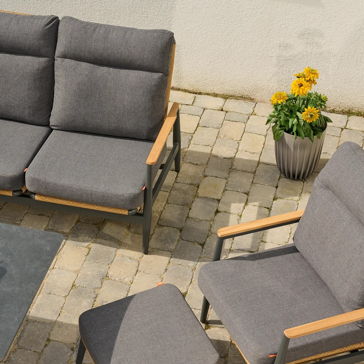Blick von oben auf ein Sofa und einen Sessel mit grauen Polstern, daneben ein Hocker, im Hintergrund gelbe Blumen in einem Pflanztopf.
