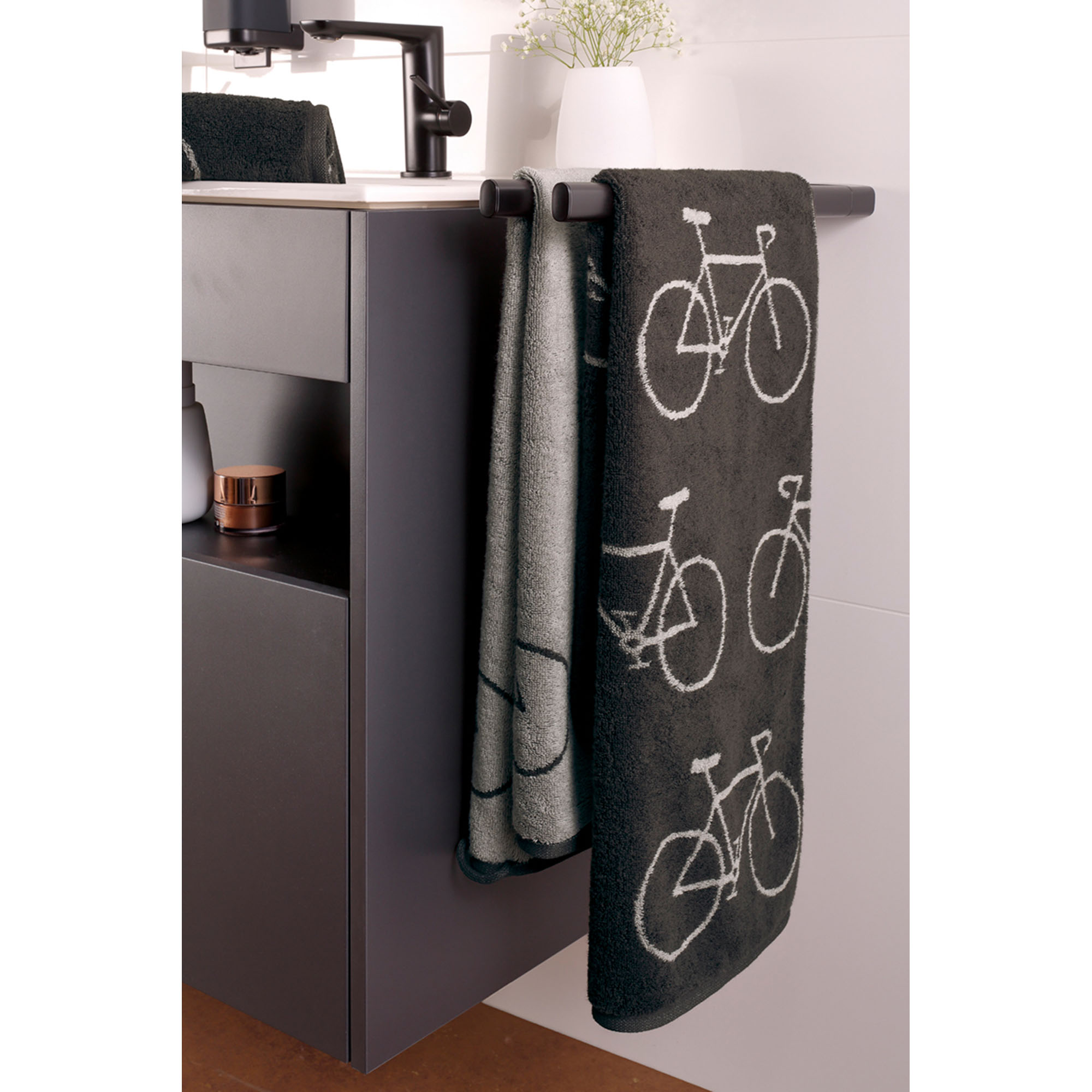 Dunkle Velo-Handtücher mit Fahrradmotiven hängen an einem grauen Badmöbel, daneben ein helleres Tuch.