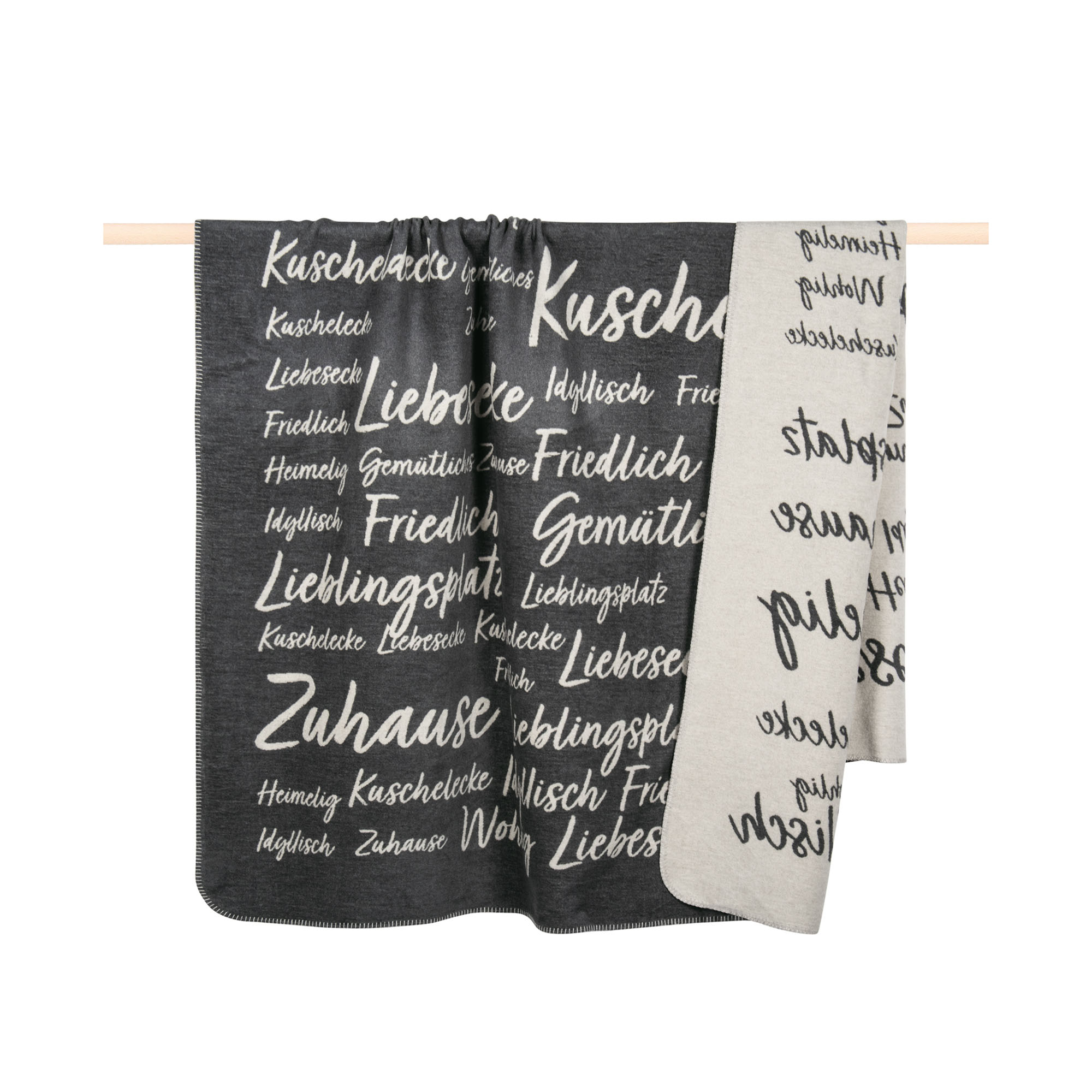 Graue Decke Snug, 150 × 200 cm, mit großflächigem schwarz-weißem Typografie-Muster, über einer Holzstange drapiert.