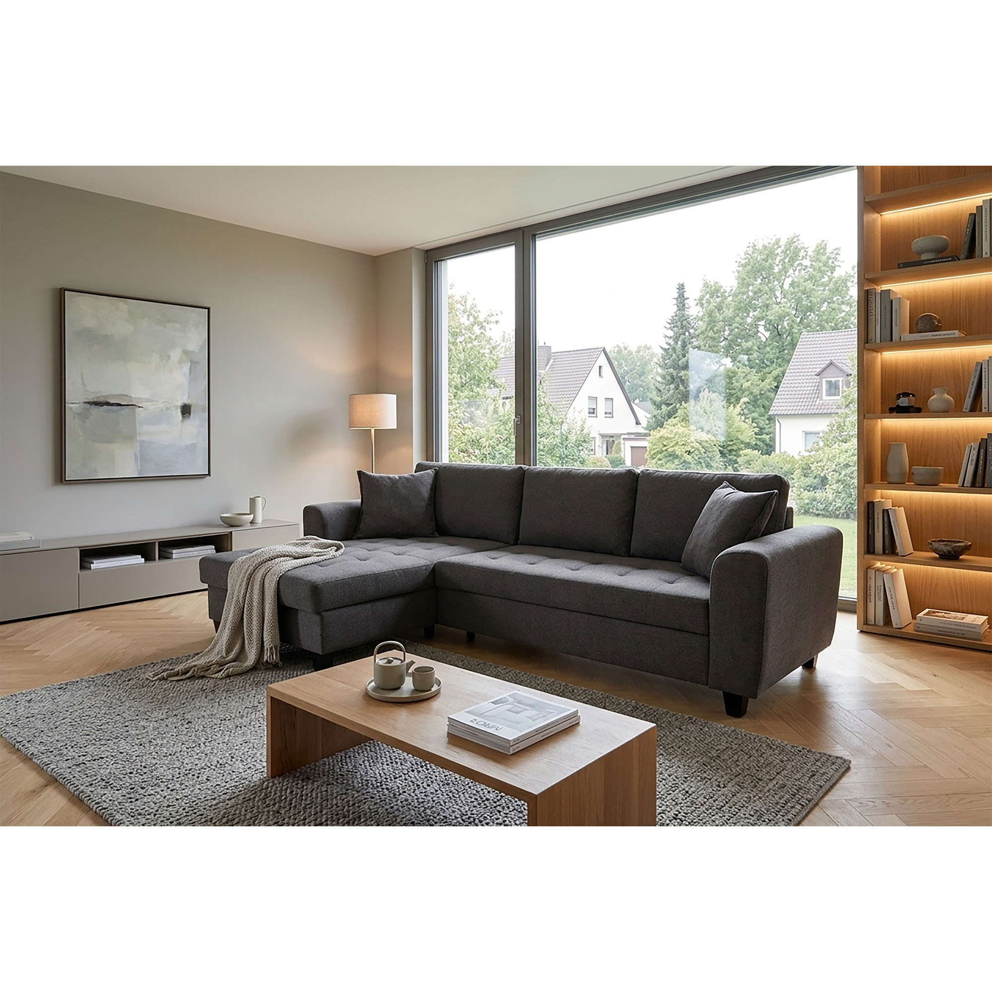 Graues Ecksofa mit links angeordneter Ottomane in einem hellen Wohnzimmer mit großem Fenster, Teppich, Couchtisch und beleuchtetem Bücherregal