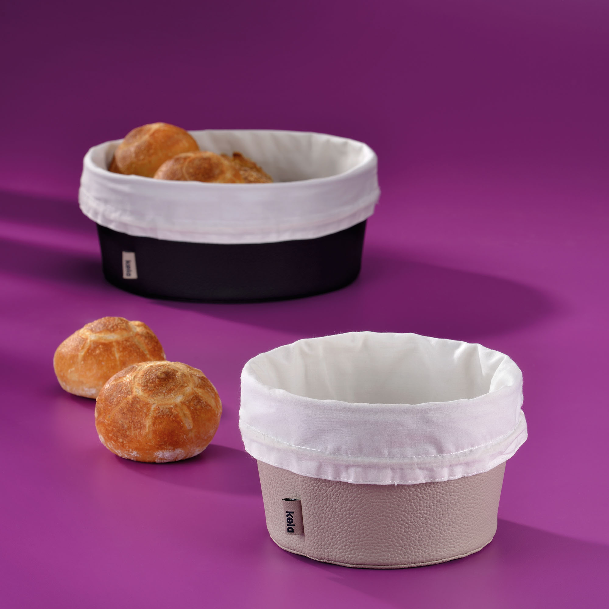 Silbergrauer und dunkler Brotkorb mit weißen Stoffeinsätzen neben mehreren Brötchen auf einer violetten Fläche